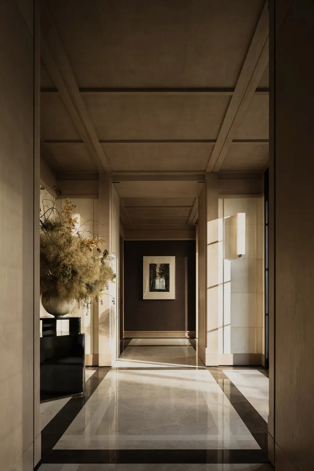 A long, minimalist luxury hallway featuring polished marble floors with dark borders, a coffered concrete ceiling, a large dried floral arrangement on a black pedestal, a framed photograph at the end of the corridor, and vertical wall sconces casting warm light.