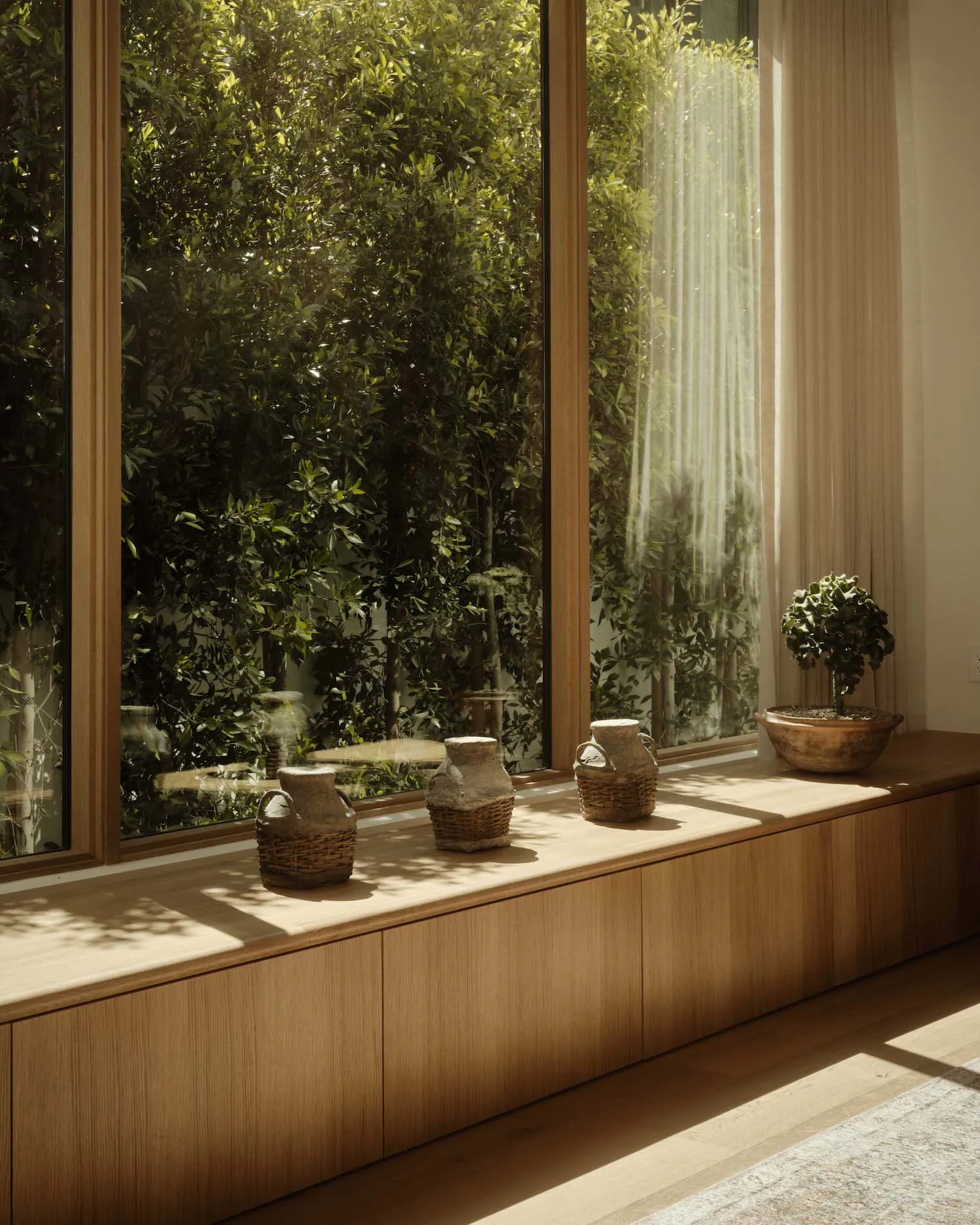 Built-in light oak window seat with large floor-to-ceiling windows overlooking dense green trees, decorated with three rustic earthenware pots in woven baskets and a bonsai tree in a wooden bowl.