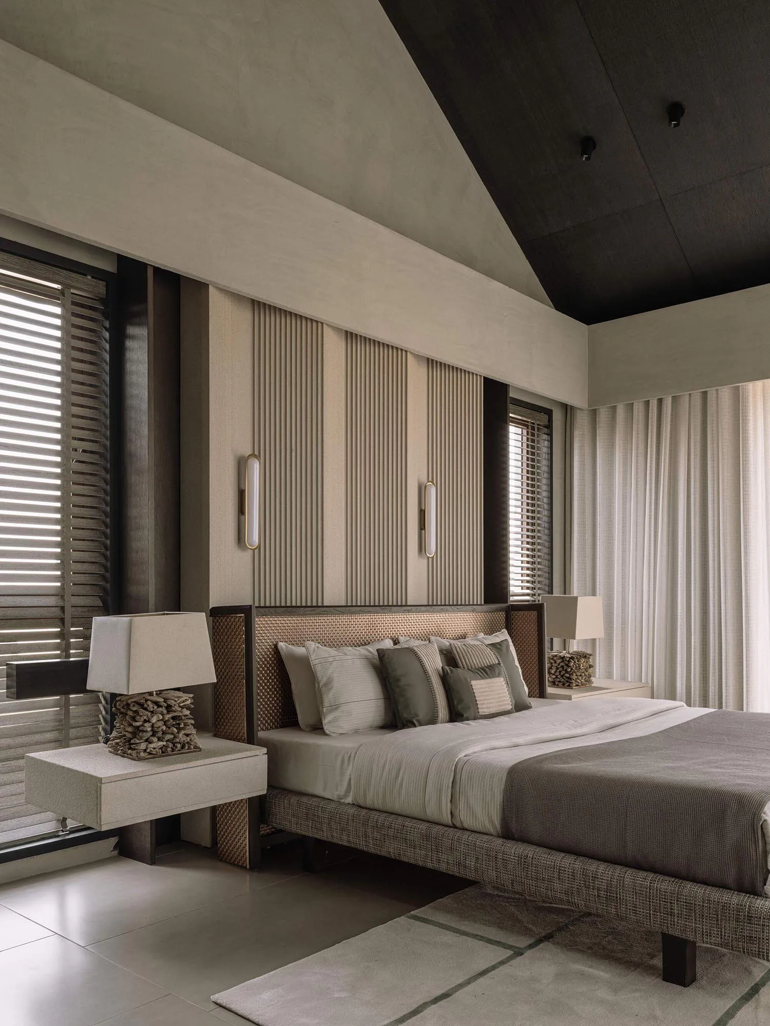 Modern luxury bedroom with tall woven rattan headboard, layered neutral bedding, floating nightstands, and dramatic angled ceiling with vertical wall panels.