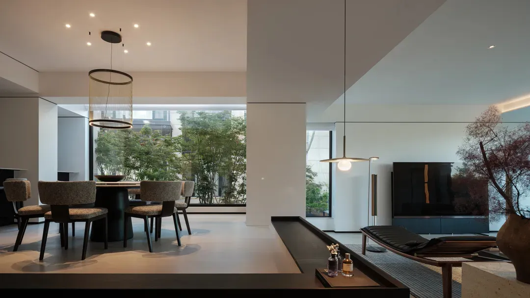 Modern minimalist open plan living and dining room featuring a round dining table with six upholstered chairs, large cylindrical pendant lights, a black leather chaise lounge, wall-mounted television, and large windows overlooking green trees.
