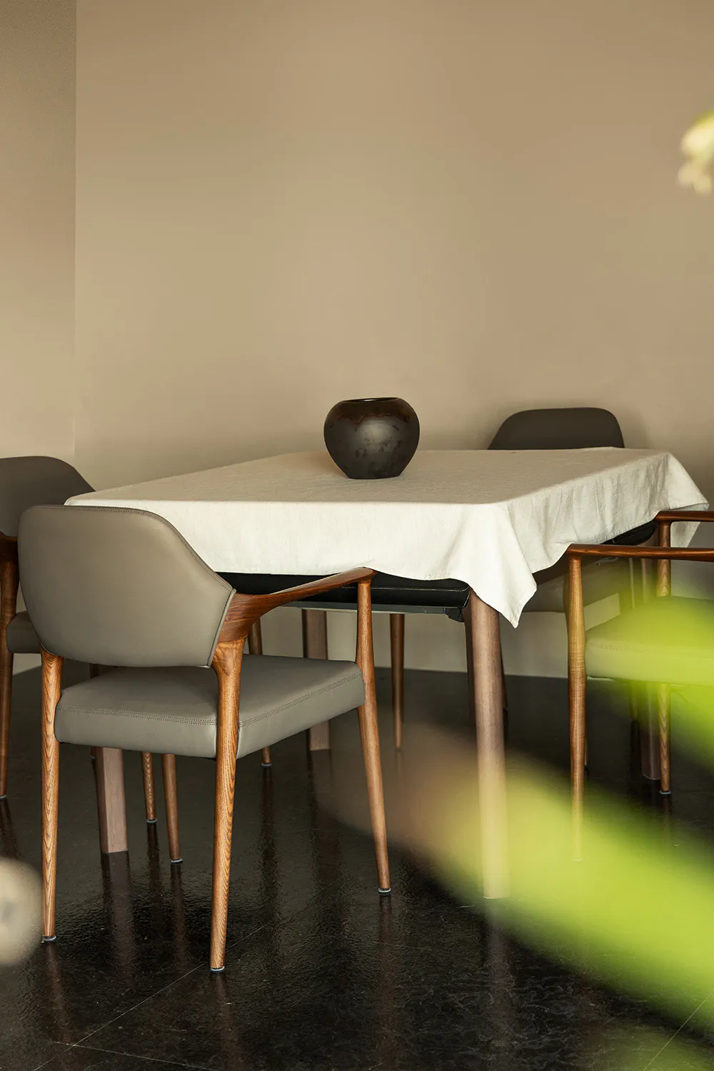 Modern minimalist dining area with walnut wood armchairs upholstered in taupe leather, a rectangular table covered with a white linen tablecloth, and a dark ceramic vase on a dark reflective floor against beige walls.