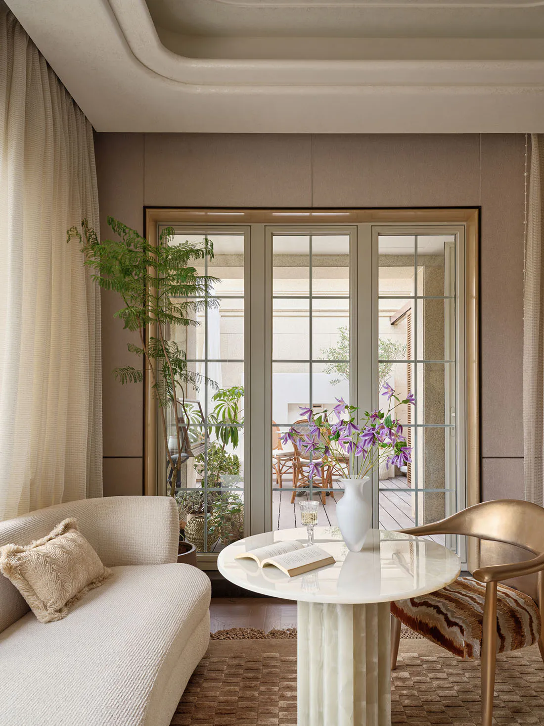 Modern living room interior featuring a curved cream sofa, a round white marble pedestal table with an open book and purple flower vase, a gold metallic chair with striped cushion, and large glass doors opening to a sunlit patio with plants.