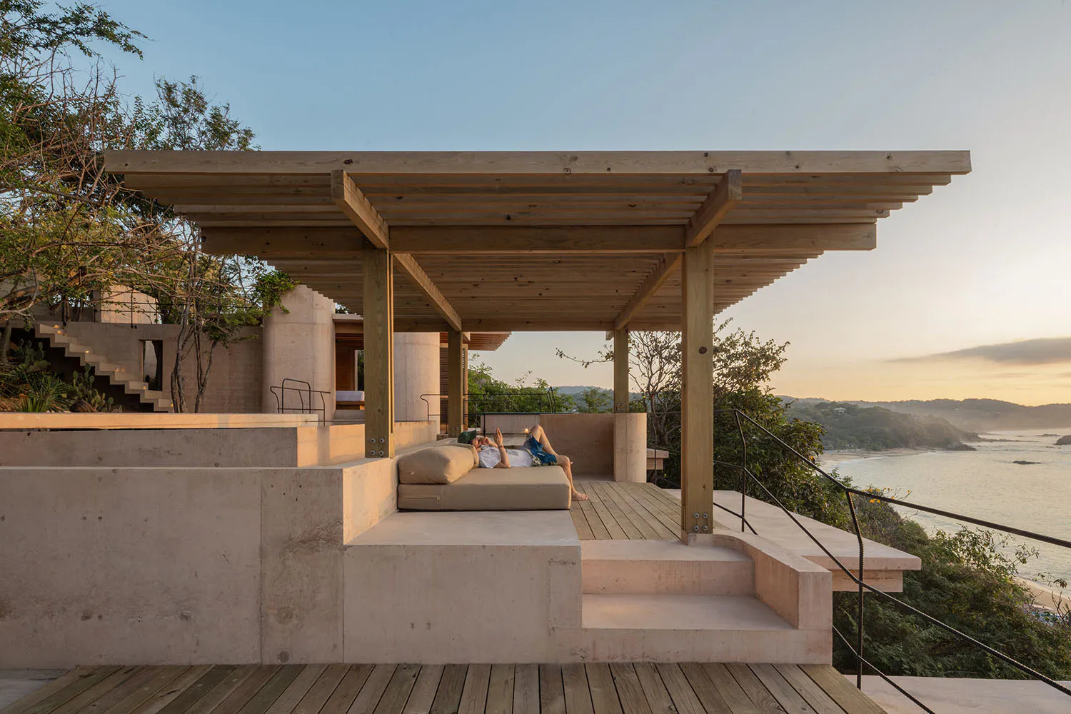 A man relaxing on a built-in daybed under a large wooden pergola on a concrete and wood ocean-view terrace at sunset.