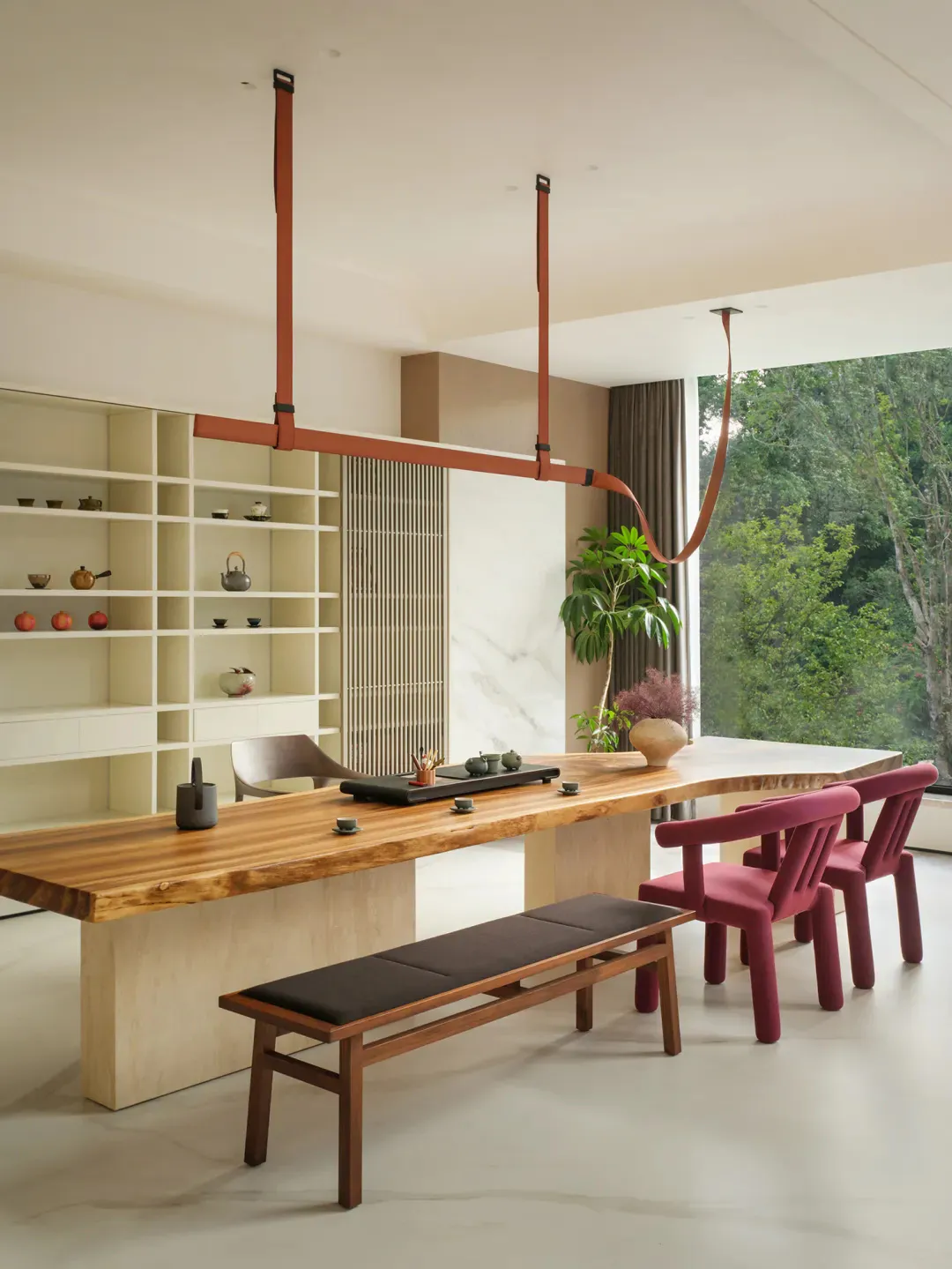 Modern dining room featuring a long live-edge walnut wood table, a suspended terracotta leather strap light fixture, three magenta upholstered chairs, a wooden bench with black cushion, built-in white shelving displaying tea ware, and large windows overlooking a lush green forest.