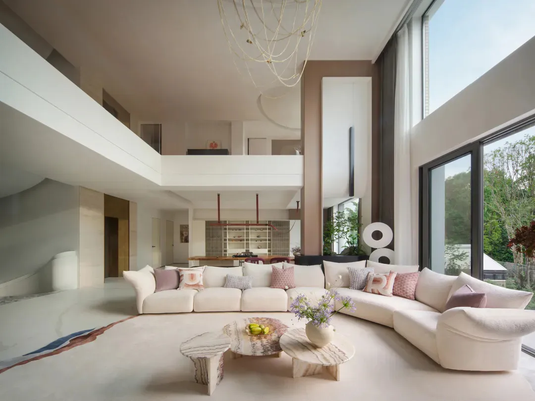 Modern double-height living room featuring a large curved white sectional sofa, three unique marble coffee tables, a sculptural rope chandelier, and floor-to-ceiling windows overlooking greenery.