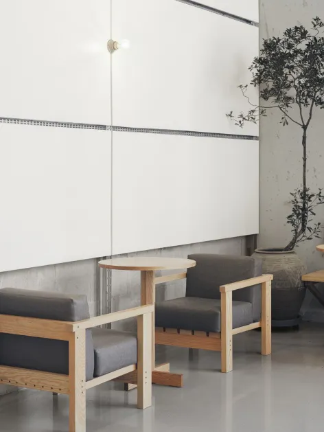 Two minimalist oak armchairs with grey upholstered cushions placed in a modern interior with white panel wall, concrete lower wall, and a large potted tree in the background.