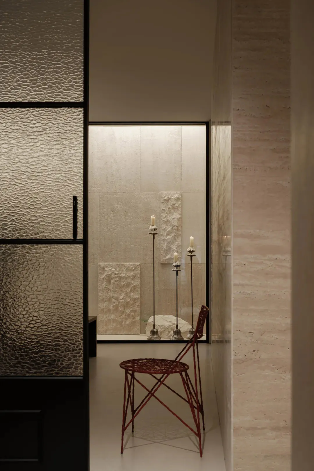 Modern minimalist hallway featuring a red woven wire chair, tall candlesticks with lit candles, and large travertine stone walls and panels.