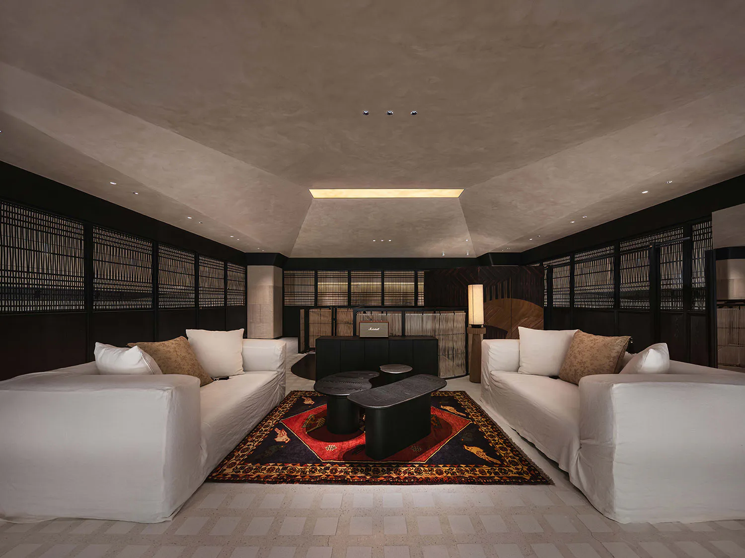 Modern Japanese-style lounge featuring two white sofas facing each other across a dark coffee table on a patterned oriental rug, with textured plaster walls and ceiling, dark wood shoji screens, and warm ambient lighting.