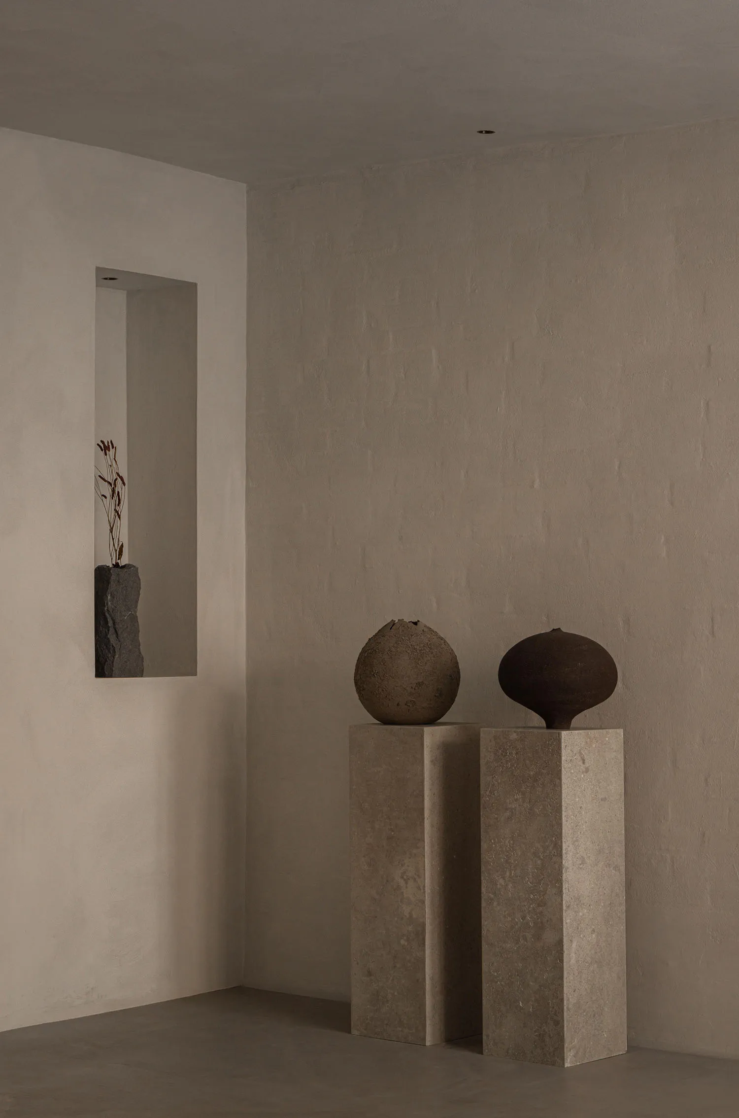 A serene minimalist interior corner with two concrete pedestals holding a textured spherical ceramic vessel and a dark organic-shaped vessel, alongside a recessed niche containing a stone vase with dried botanicals.