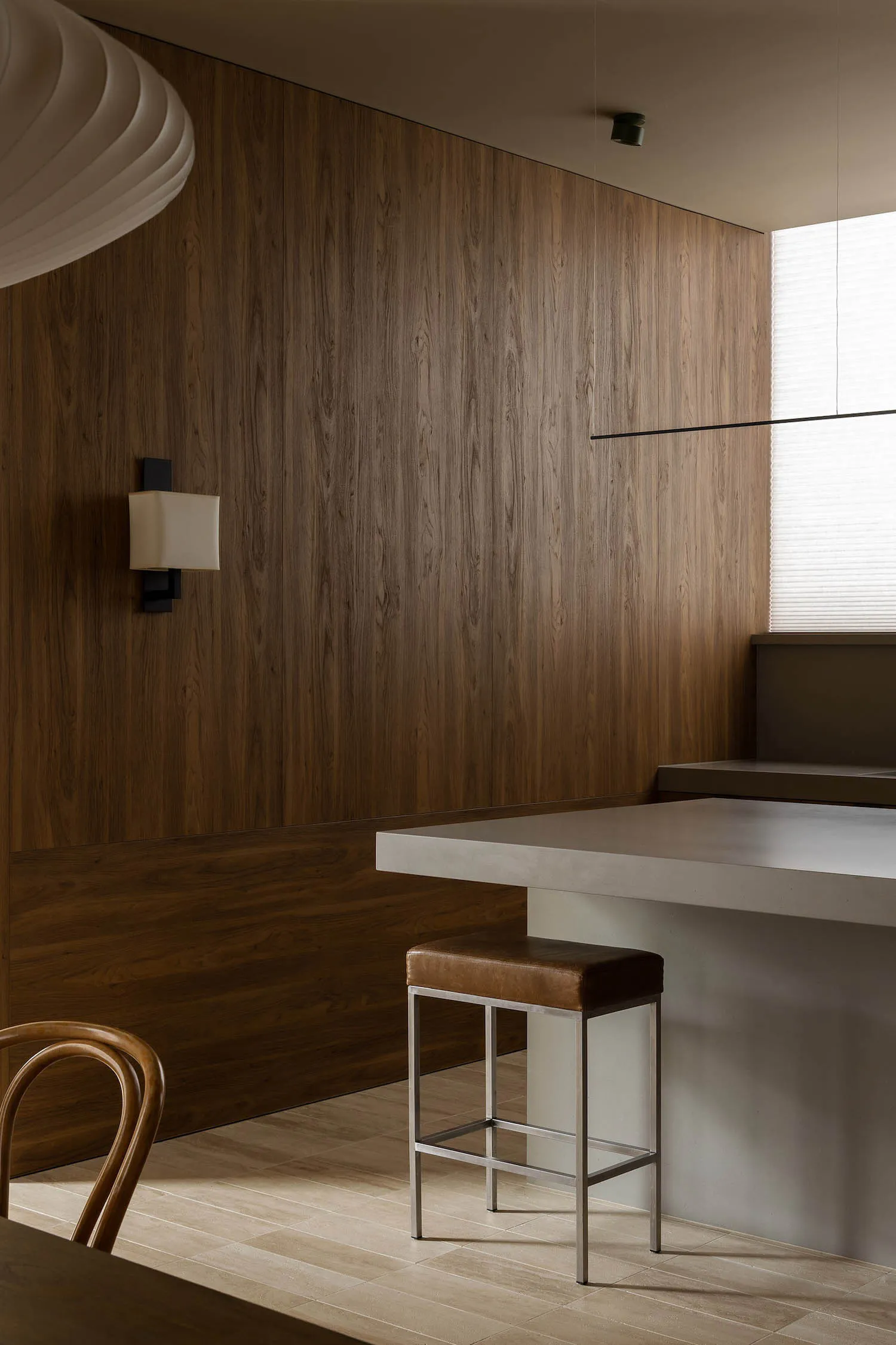 Modern minimalist kitchen featuring a dark walnut wood paneled wall, a concrete kitchen island, a brown leather bar stool with metal frame, and a sculptural white pendant light.
