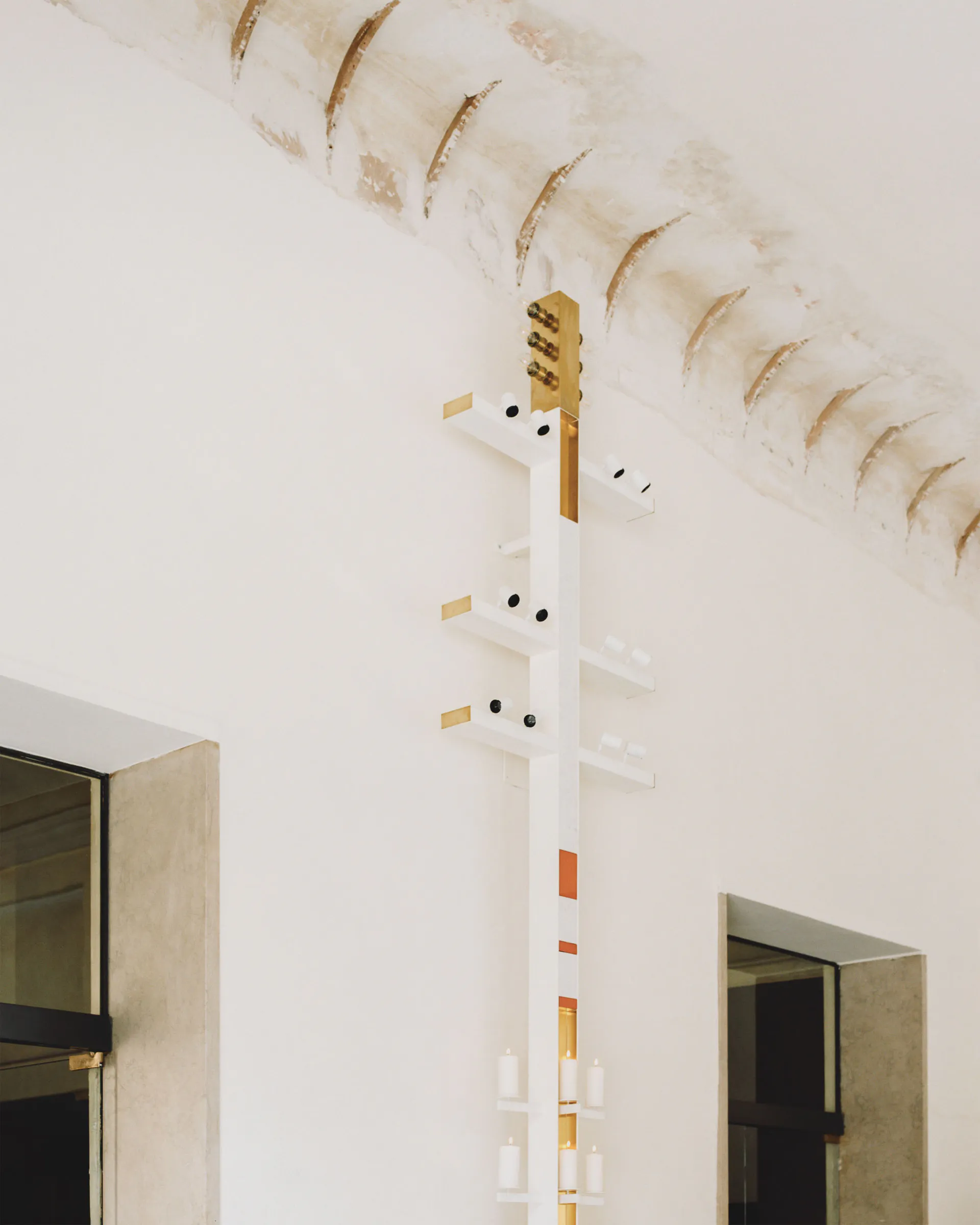 Vertical sculptural wall light fixture made of white shelves, brass accents, and electric candles mounted on a white plaster wall under a curved, aged vaulted ceiling with two doorways visible on either side.