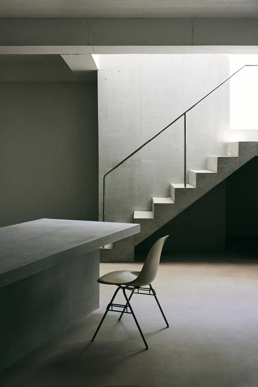 Minimalist brutalist interior with raw concrete walls, a floating concrete staircase with thin metal handrail, and a beige Eames-style chair placed next to a concrete counter.