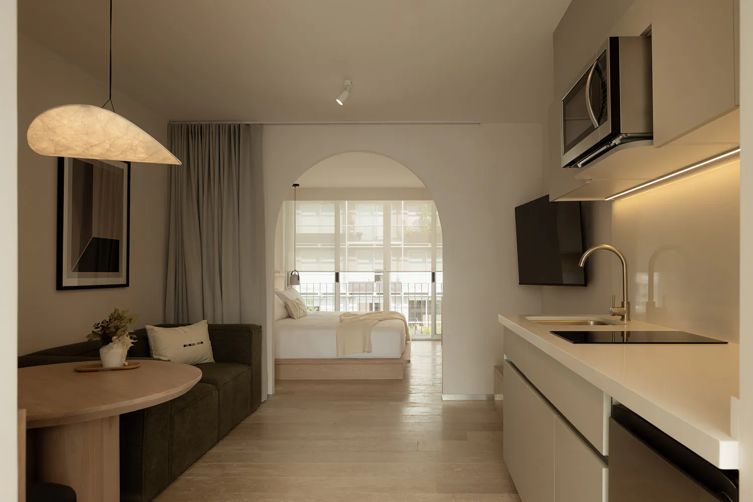 Modern minimalist studio apartment interior with white arched doorway leading to bedroom, green sofa, round wooden table, and compact kitchen with built-in microwave and induction cooktop.