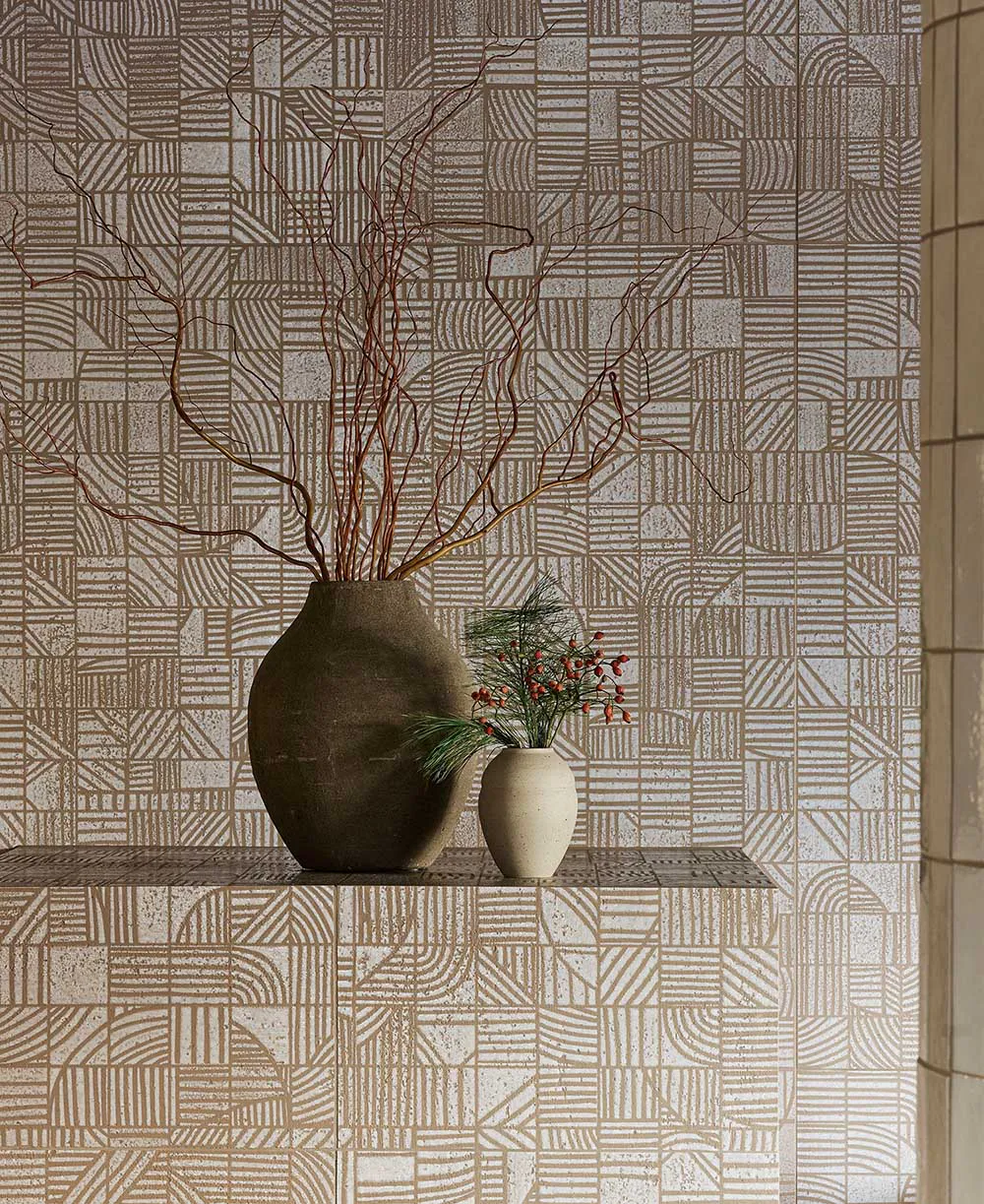 Still life arrangement on a textured shelf featuring a large dark earthenware vase with twisted brown branches and a small off-white ceramic vase with pine and red berries, set against a wall of beige and white geometric patterned tiles.