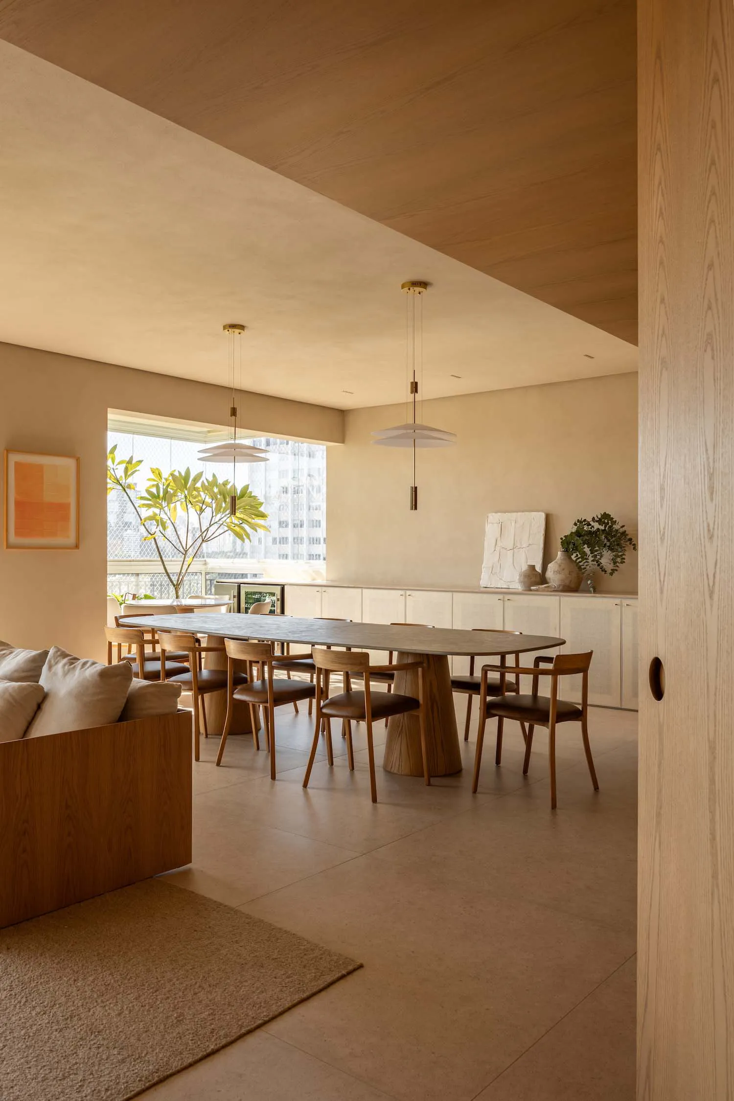 Modern minimalist dining room featuring a large oval travertine-topped oak dining table surrounded by wooden chairs, two white pendant lights, built-in oak cabinetry, and a large window overlooking city buildings.