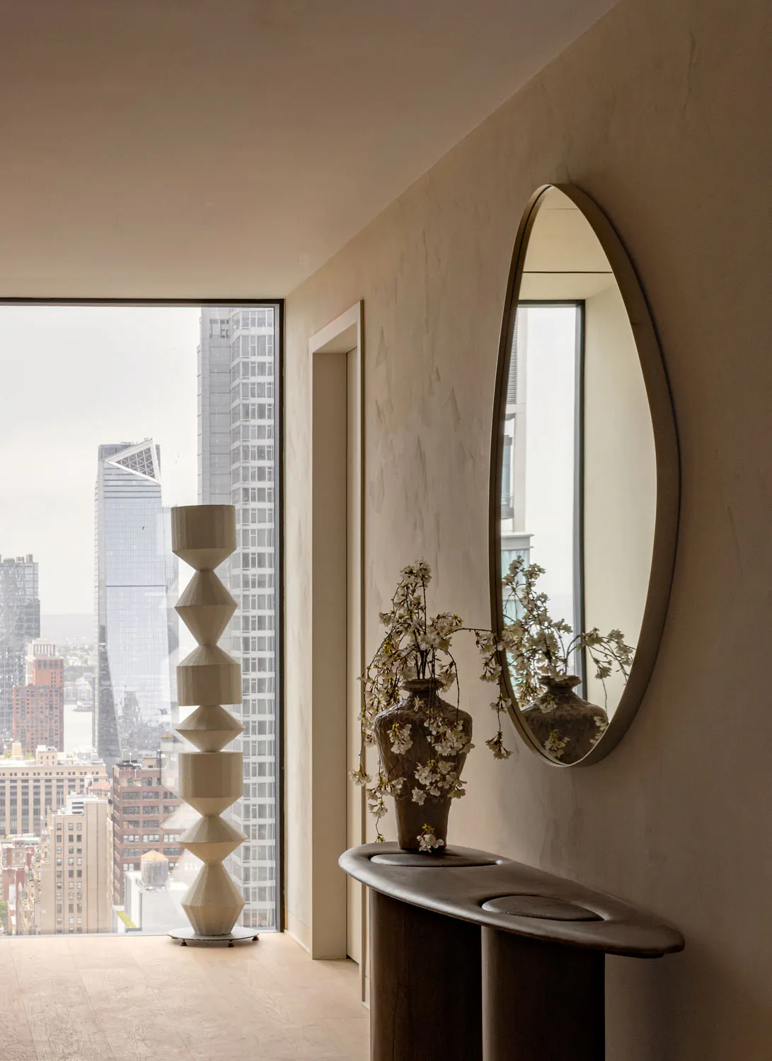 Modern minimalist entryway in a New York high-rise apartment featuring a tall sculptural beige floor lamp, an oval mirror, a dark organic-shaped travertine console table with a vase of white cherry blossoms, and a panoramic city view through large windows.