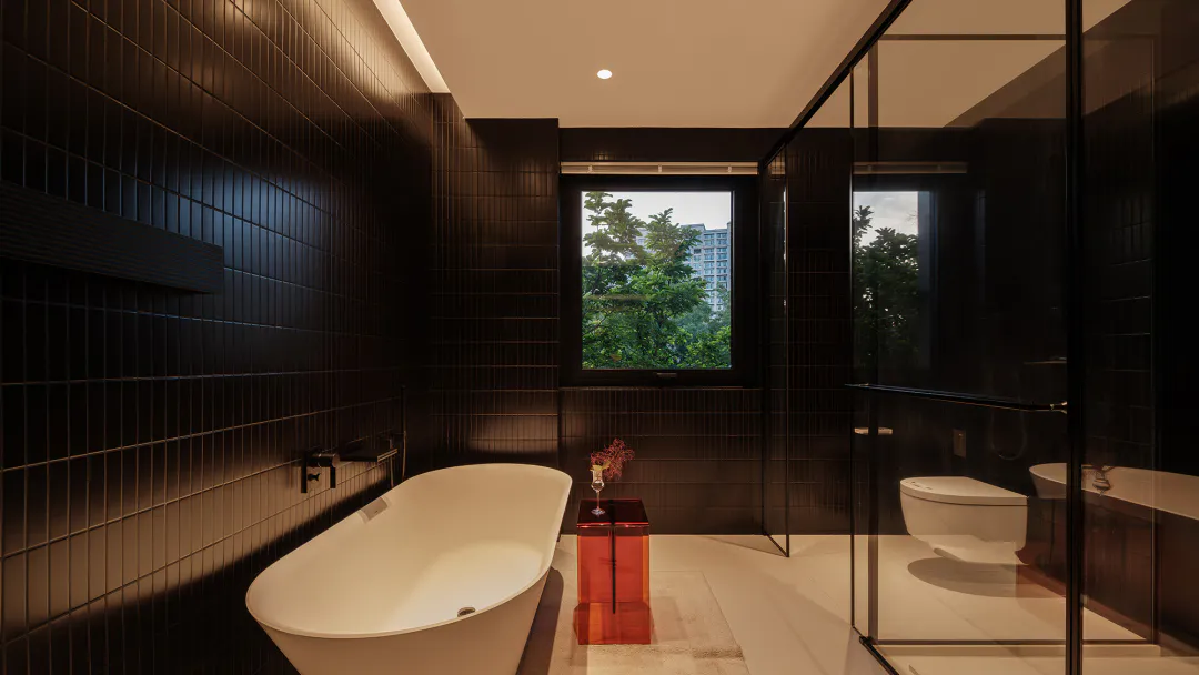 Modern dark-themed bathroom interior with a white freestanding bathtub, black rectangular wall tiles, glass-enclosed shower and toilet area, and a small red side table with flowers next to a large window overlooking green trees and buildings.