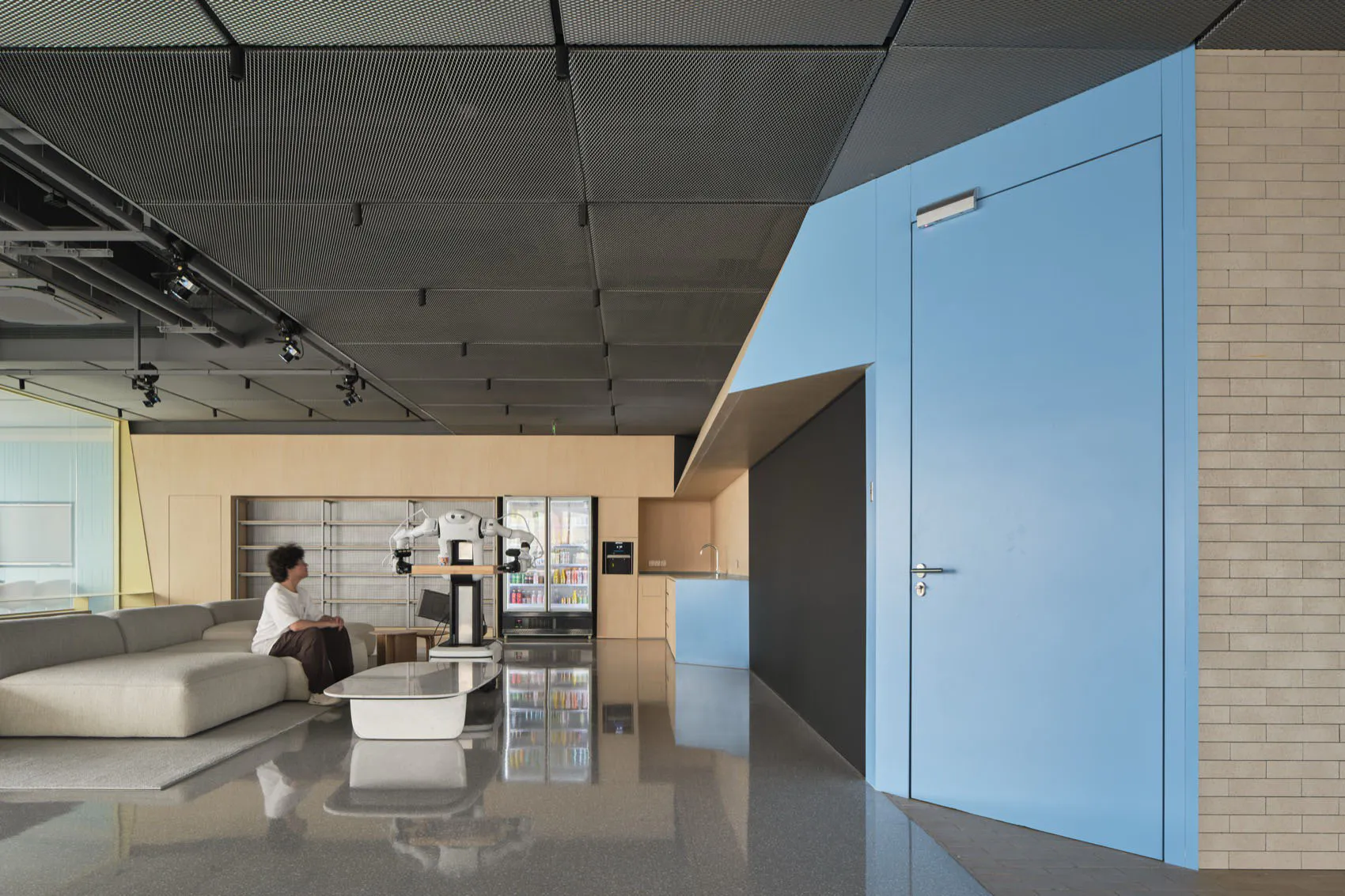 Modern open-plan office lounge featuring a large white robotic arm, reflective polished concrete floor, light gray sectional sofa, blue door, and beverage refrigerators.