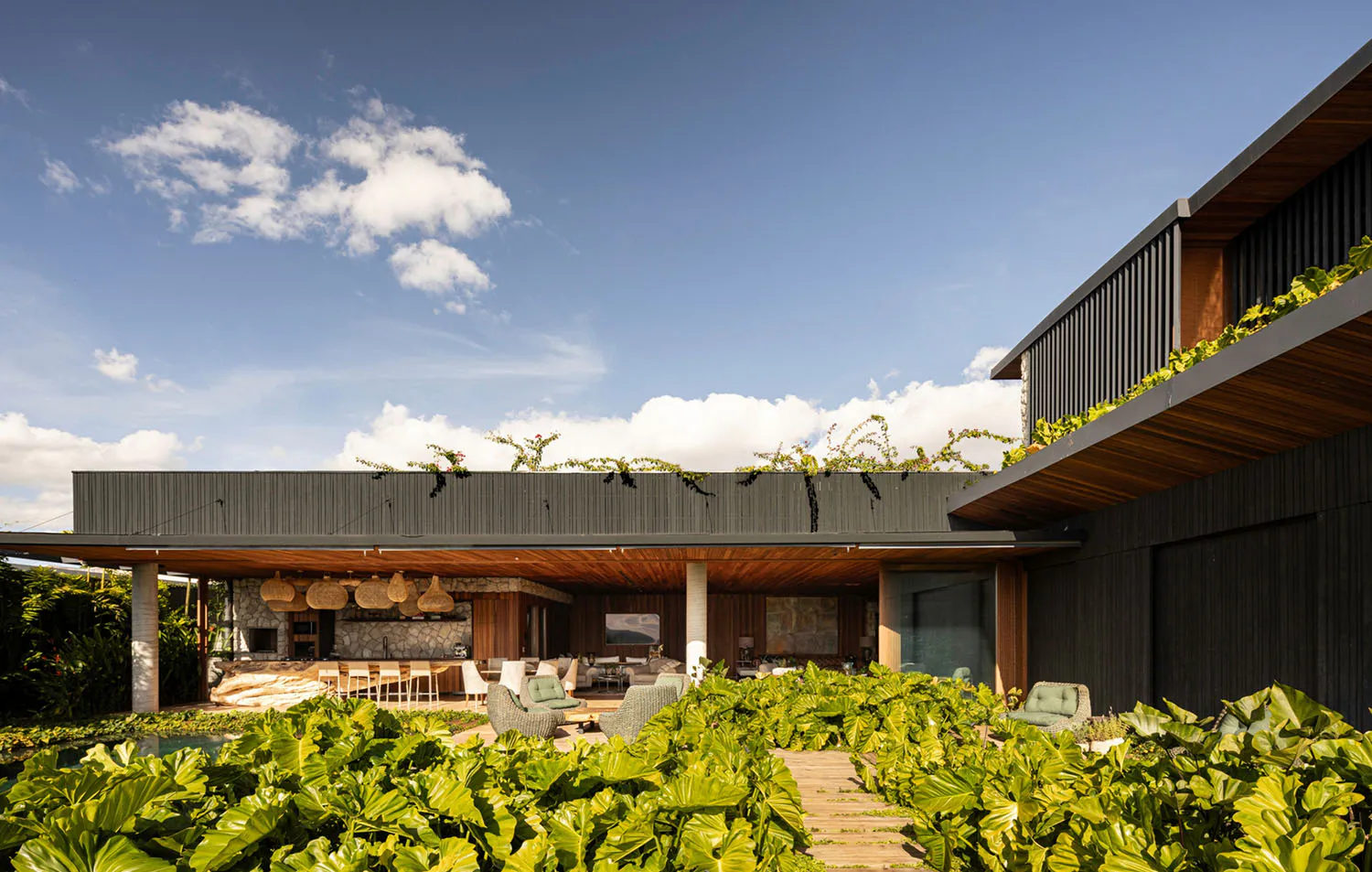Modern tropical house exterior featuring a large covered outdoor patio with wooden ceiling, woven pendant lights, stone walls, and lush green tropical plants in the foreground under a bright blue sky.