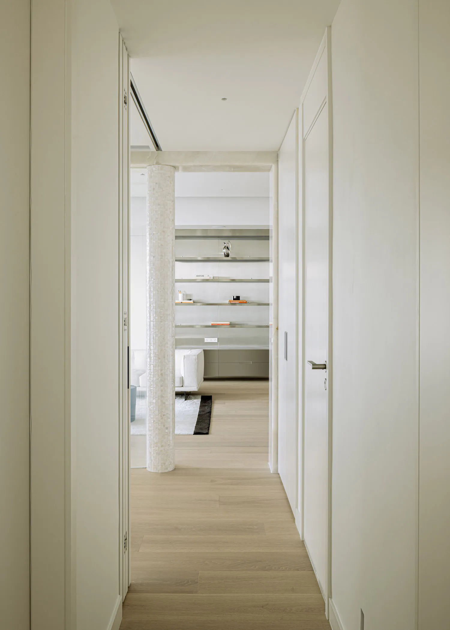 View from a narrow white hallway looking into a modern study area featuring a cylindrical mother-of-pearl column, built-in floating shelves with decorative objects, light wood flooring, and a white sofa in the background.