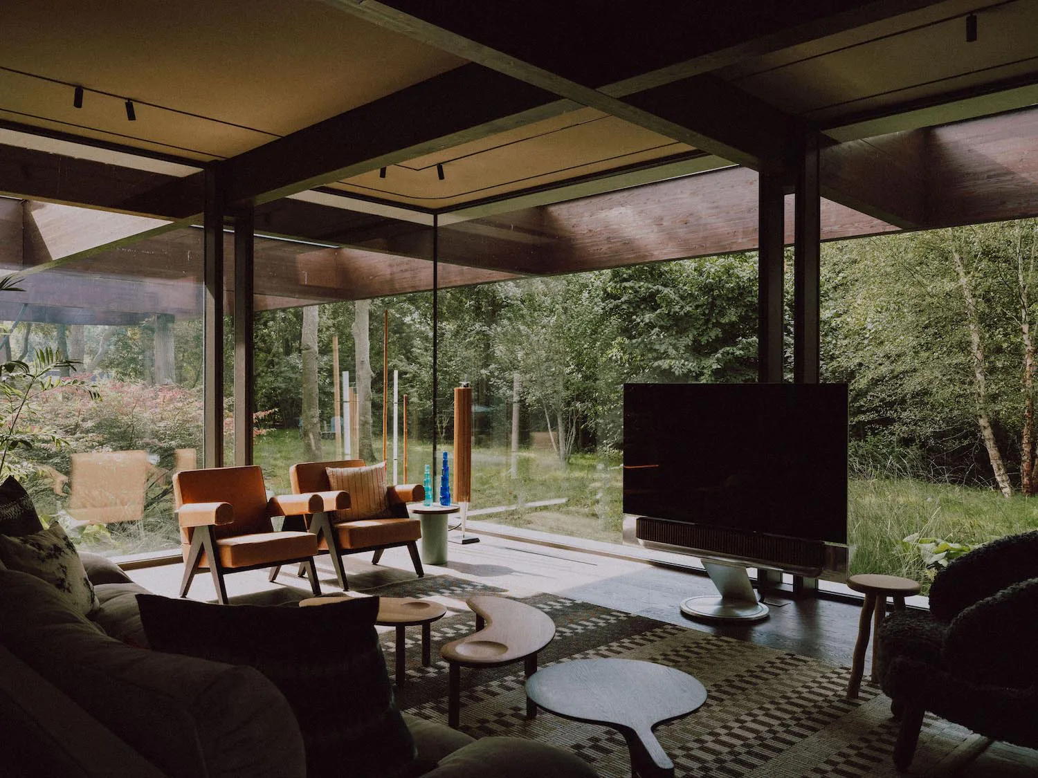 Modern living room interior featuring large glass walls overlooking a forest, orange leather armchairs, wooden ceiling beams, a large flat-screen TV, and a patterned area rug with organic-shaped coffee tables.