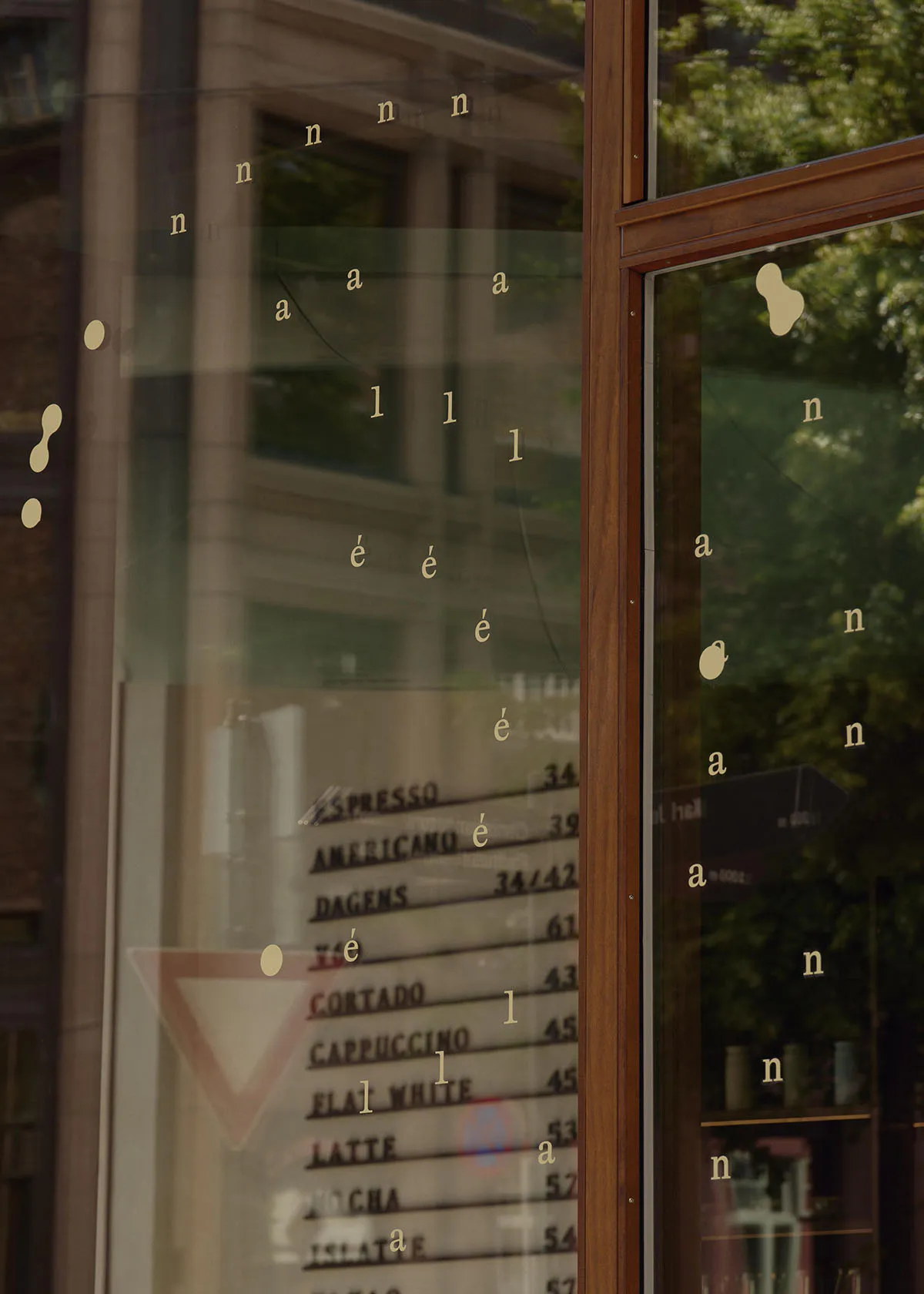 Close-up view of a modern cafe's large glass window featuring gold vinyl lettering spelling coffee-related words, a wooden frame, and reflections of green trees and a neighboring building.