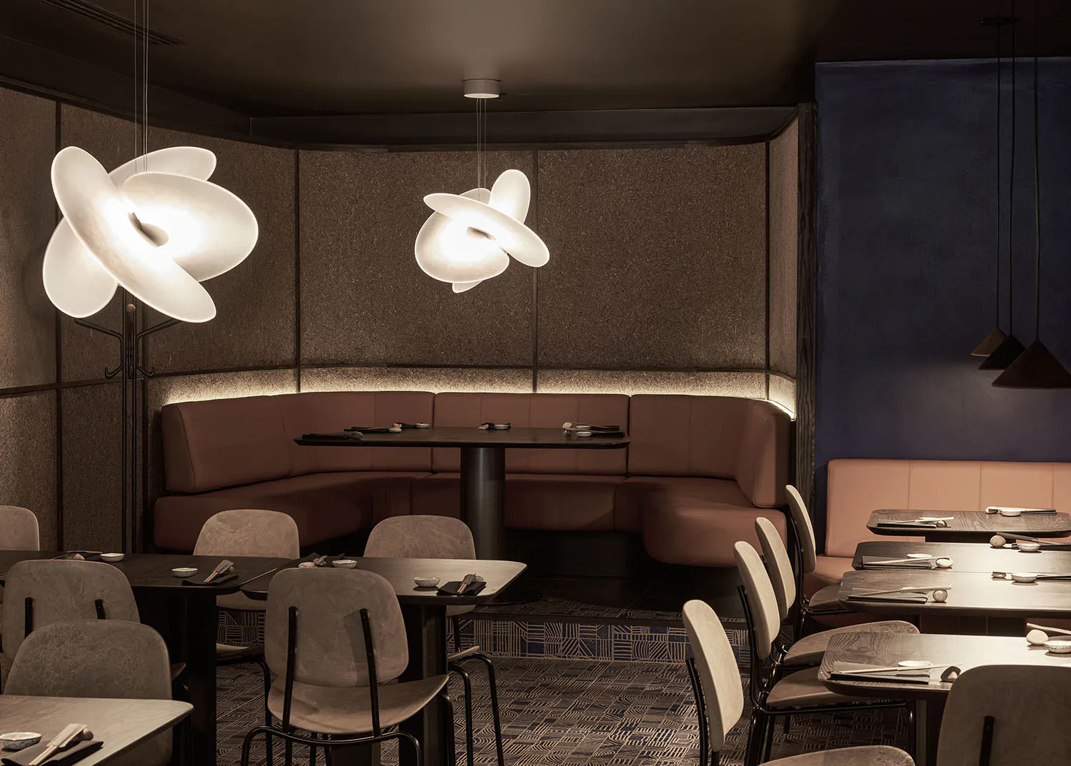 Modern Japanese-inspired restaurant interior featuring curved brown leather banquettes, dark textured walls, sculptural white flower-shaped pendant lights, and neatly set dining tables with grey upholstered chairs.