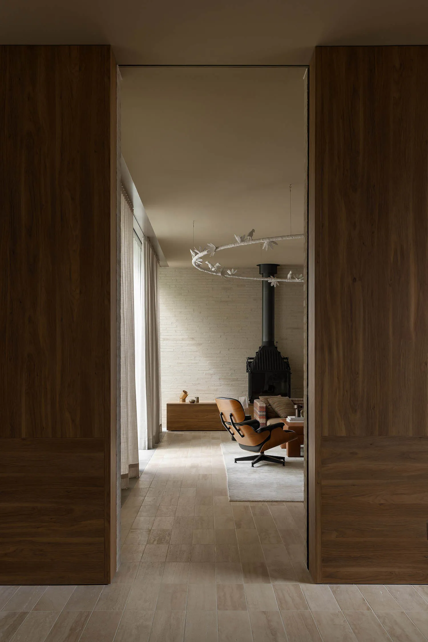 View through dark walnut wood sliding doors into a modern minimalist living room featuring a sculptural white bird chandelier, black wood-burning stove, brown leather Eames lounge chair, and light wood flooring.