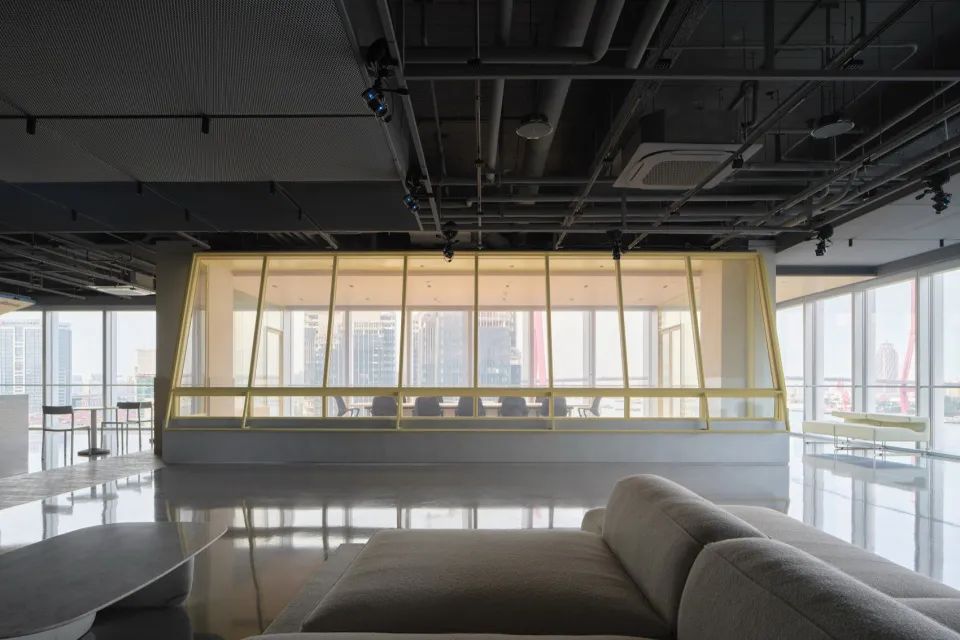Modern open-plan office lounge with gray sectional sofa in foreground, glossy reflective floor, and a prominent yellow-framed glass meeting room in the background with city buildings visible through large windows.