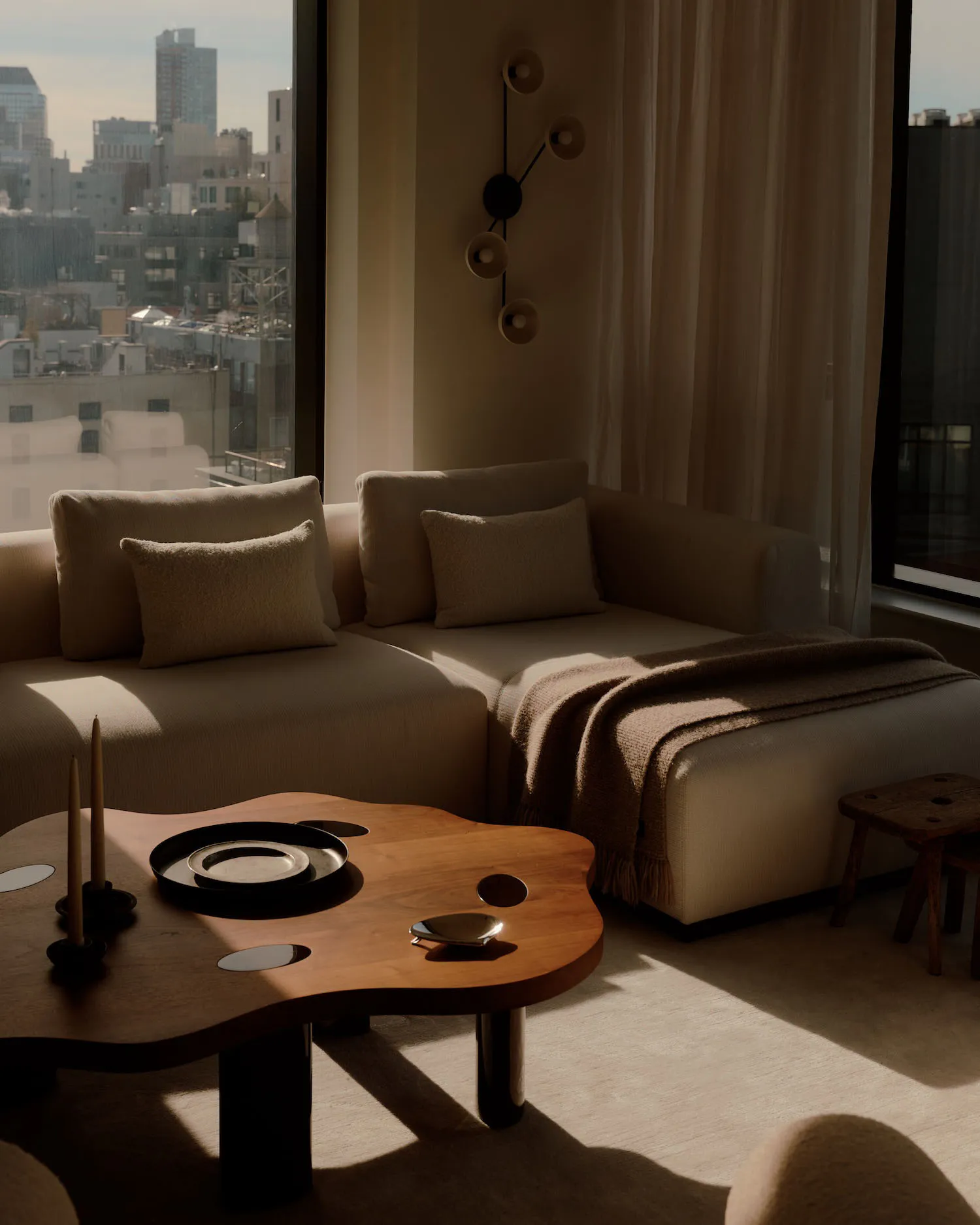 A modern minimalist living room featuring a beige L-shaped sectional sofa, a sculptural walnut amoeba-shaped coffee table with black trays and candle holders, a brown throw blanket, and a unique wall sconce, with large windows showing a city view.