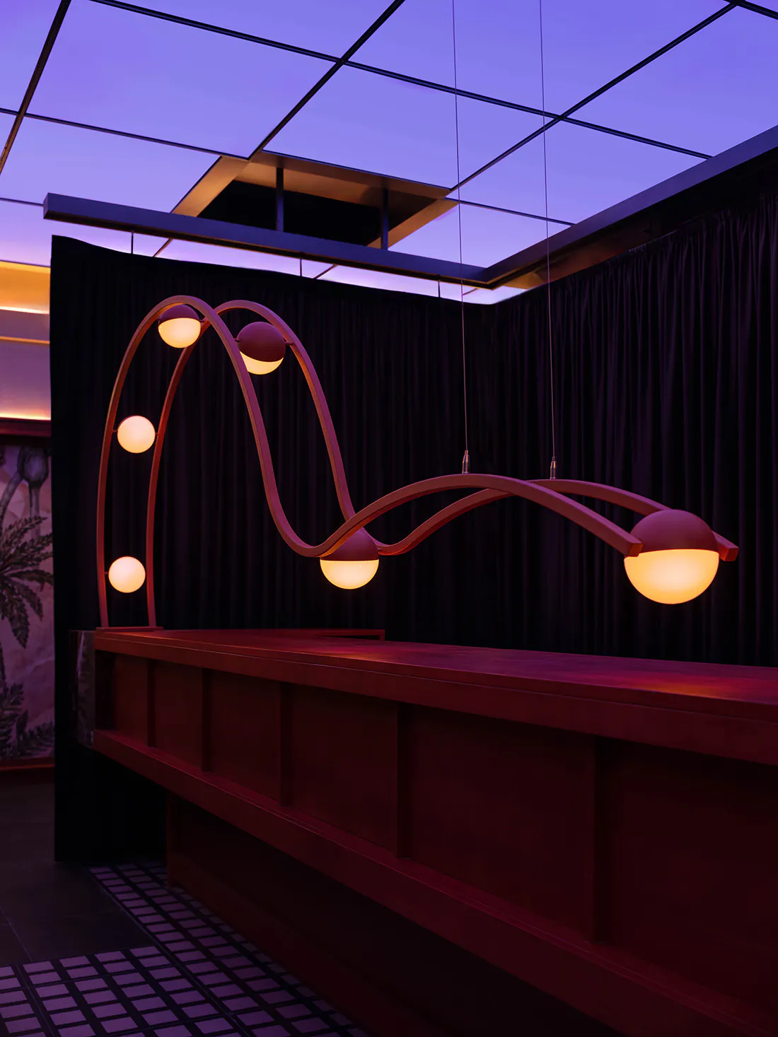 Modern reception desk made of walnut wood with a large, artistic, wavy pendant light fixture featuring multiple glowing spherical lamps against dark curtains and a purple ceiling.