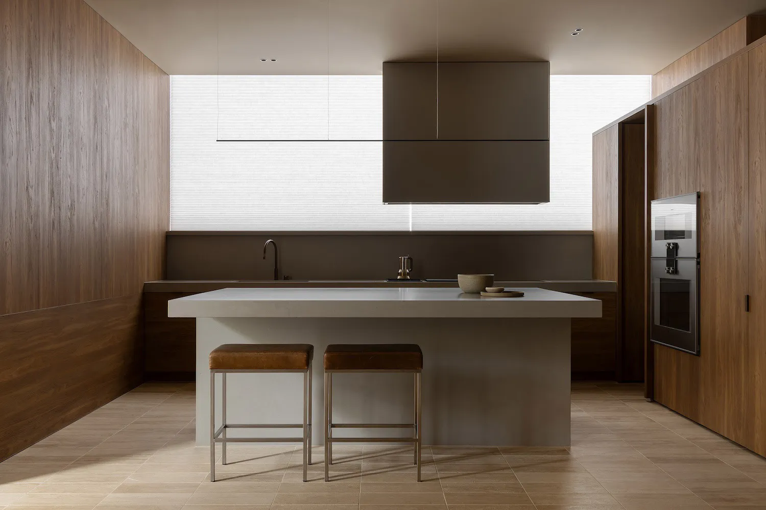 Modern minimalist kitchen interior with a large concrete central island, two brown leather bar stools, walnut wood walls and cabinetry, and a large window with sheer blinds in the background.