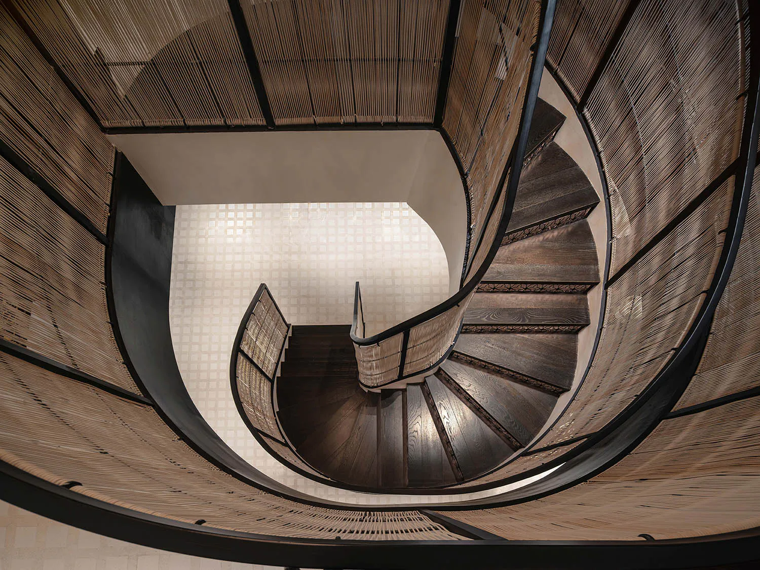 Looking down at a dramatic dark wood spiral staircase with intricate bamboo screen railings and walls, featuring a light-colored tiled floor visible at the bottom.