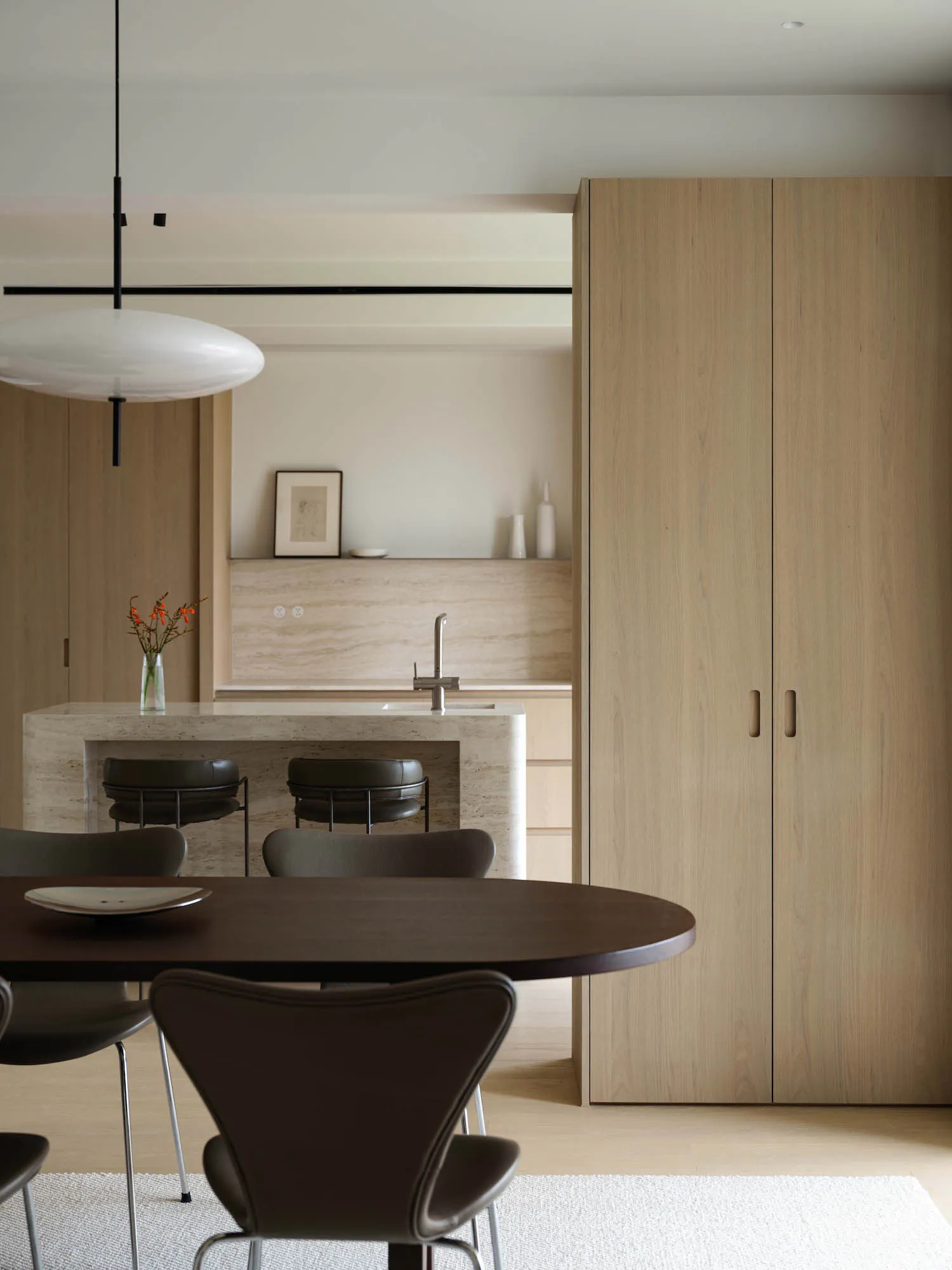 Modern minimalist dining room with dark brown dining table and chairs, travertine kitchen island with two bar stools, large light wood wardrobe, and white pendant lamp.