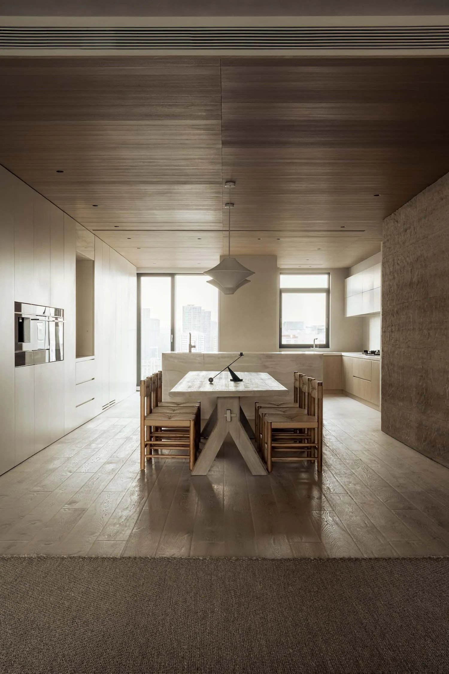 Modern minimalist dining room featuring a large oak trestle table with wooden chairs, a geometric white pendant lamp, light wood flooring, and floor-to-ceiling windows overlooking a city skyline.