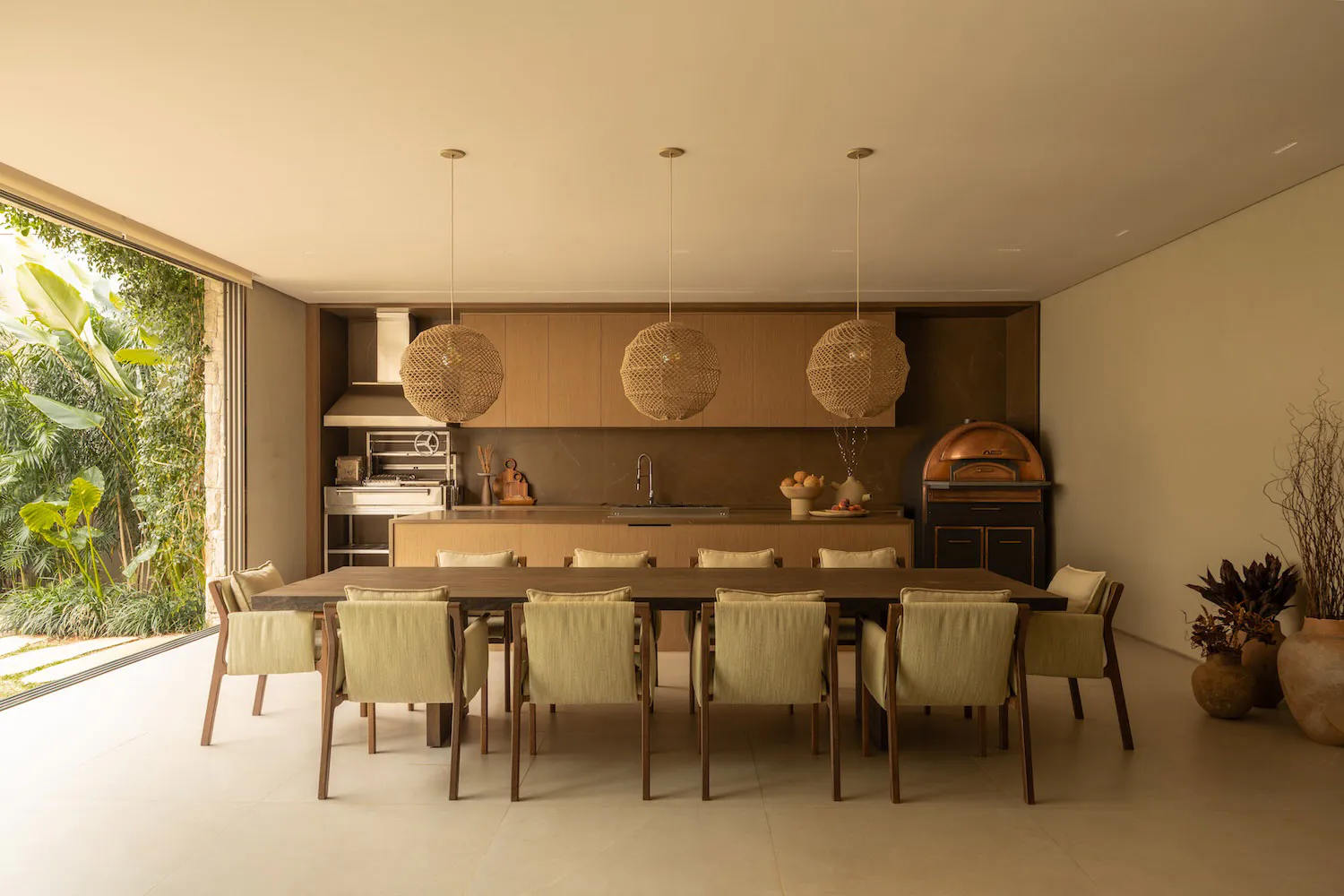 Modern open-plan dining room with a long dark wood table, ten upholstered chairs, three large woven rattan pendant lights, a built-in kitchen with wooden cabinetry, and a copper pizza oven visible in the background, opening to a lush tropical garden.