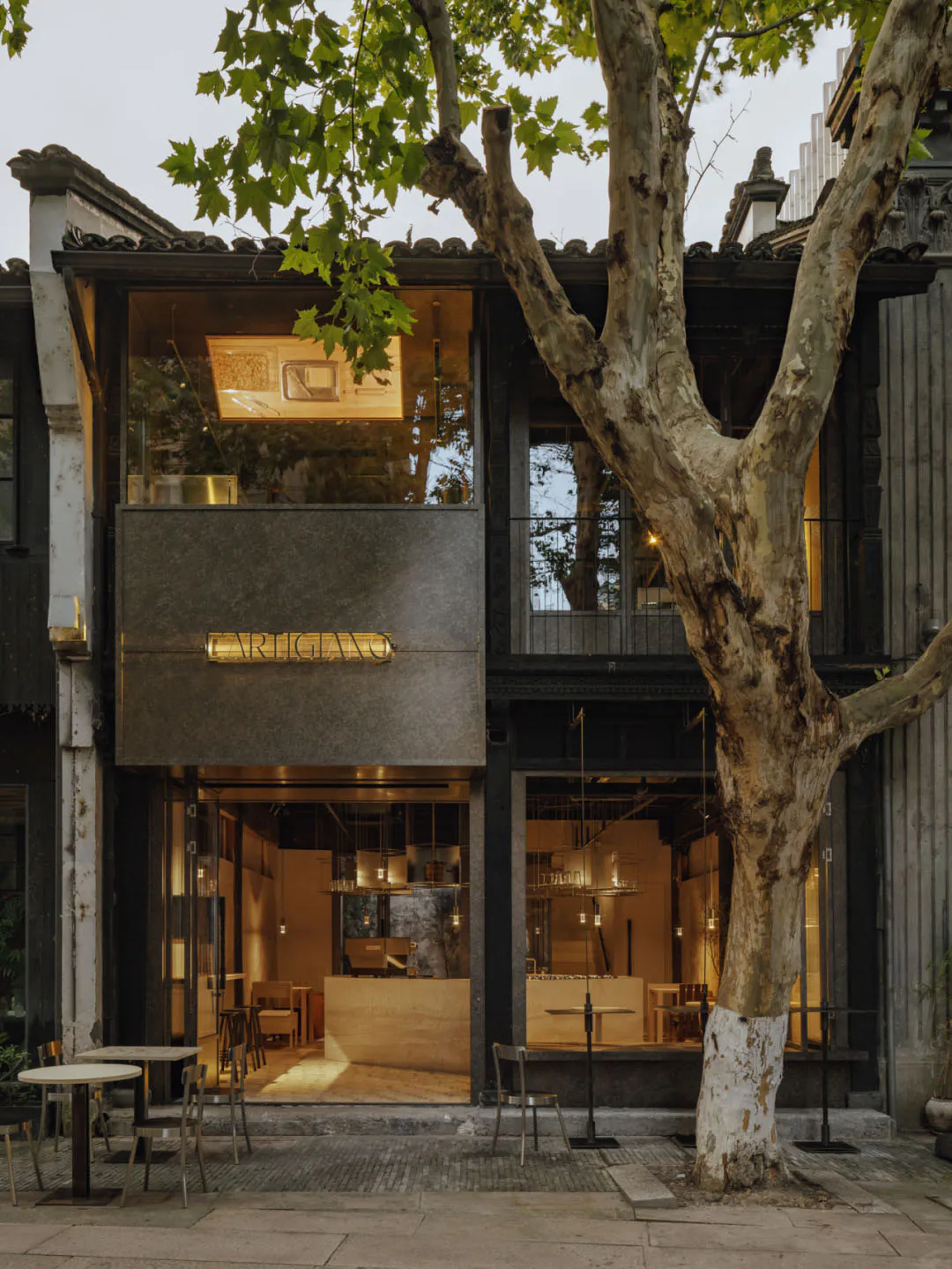 Street view of a modern cafe named Carigiano with large glass windows, concrete upper facade, warm interior lighting, outdoor seating, and a large tree with whitewashed trunk in front of traditional Chinese architecture.