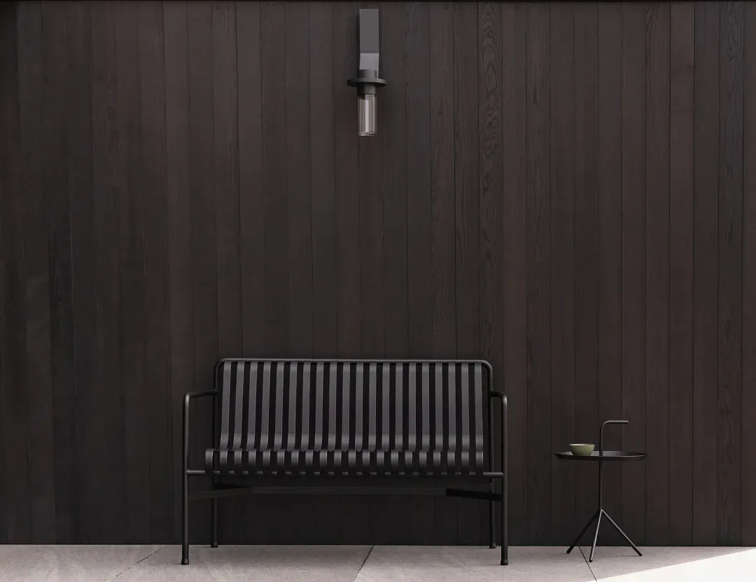 Modern black metal outdoor sofa with vertical slats and a small round side table holding a green cup, placed against a dark vertical wood-paneled wall with a black wall sconce above.