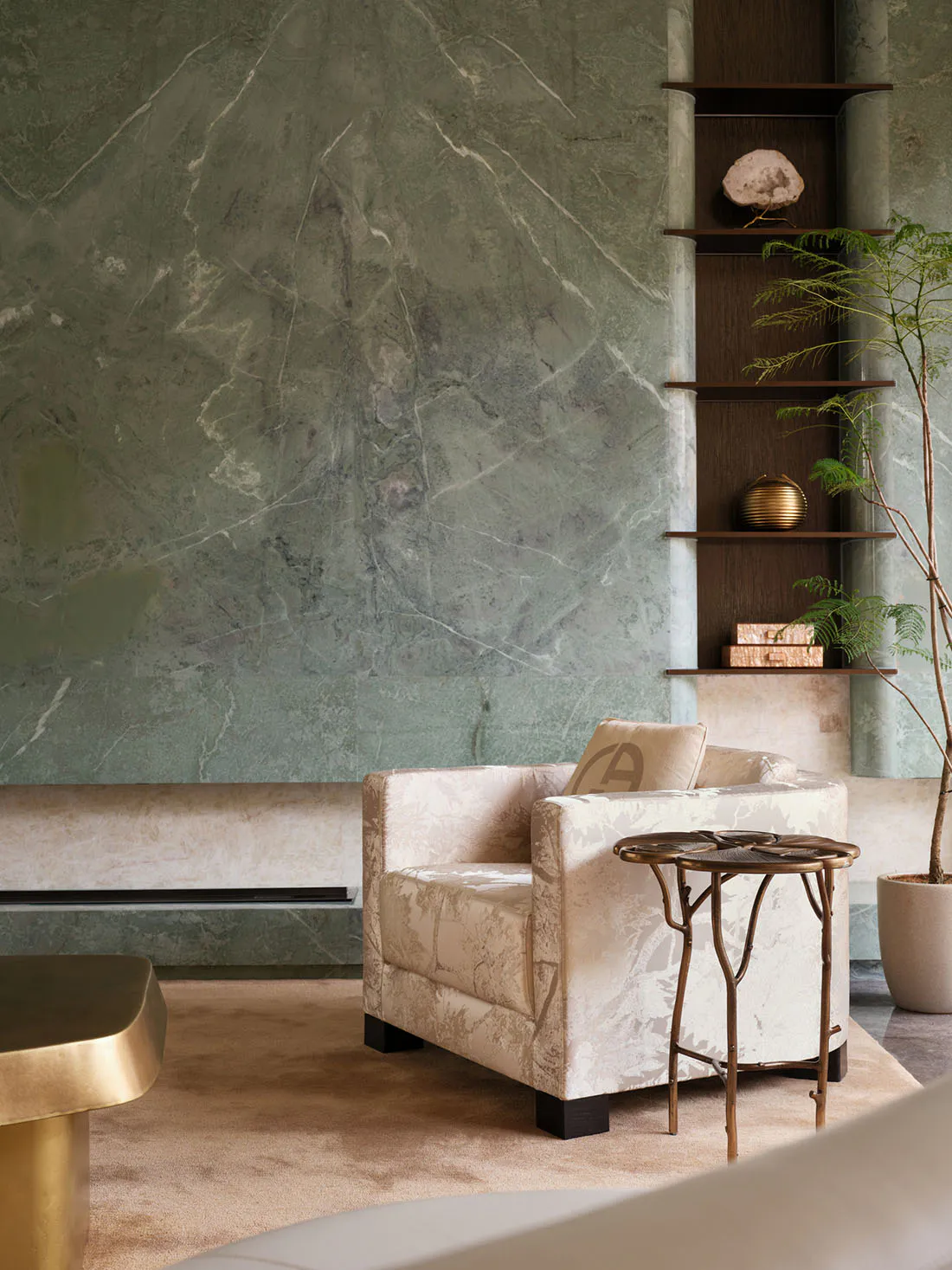 Modern living room interior with sage green marble wall, built-in wooden shelving displaying decorative objects, a patterned cream armchair with monogram pillow, and an organic-shaped brass side table on a beige carpet.