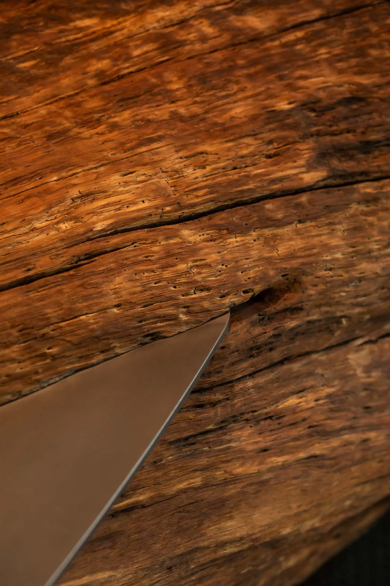 Close-up photograph of a metallic knife blade inserted into a dark, weathered walnut wooden surface with prominent grain and cracks.