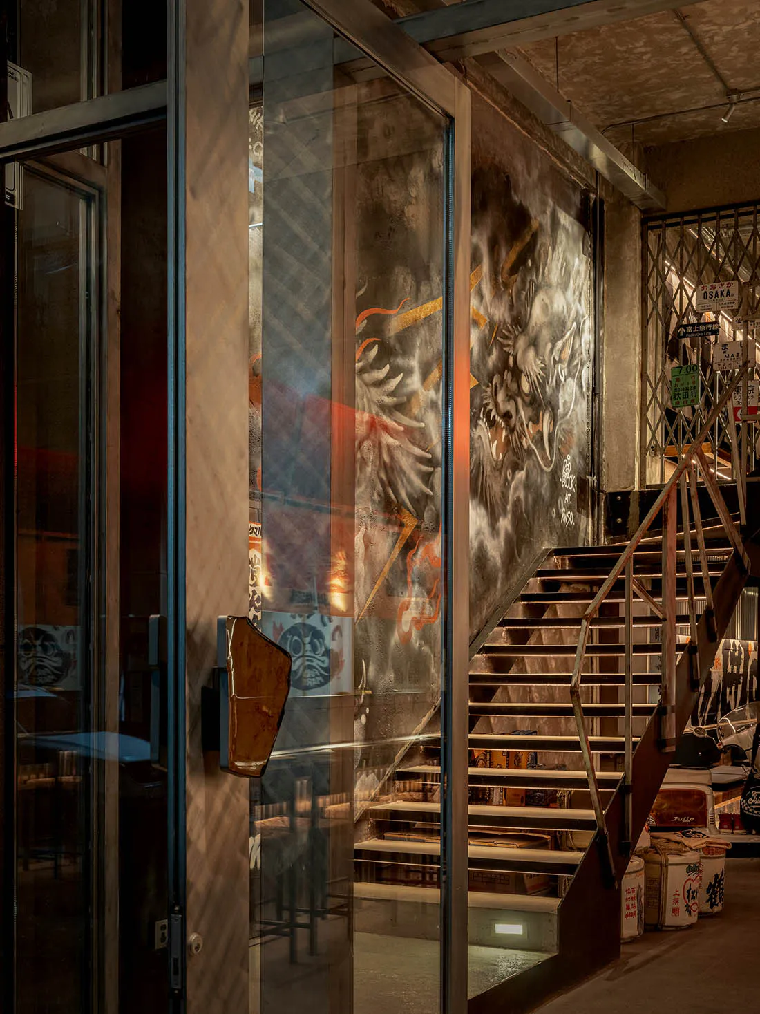 Interior view of an industrial staircase with a large dramatic dragon mural painted on exposed concrete wall, metal railings, and glass partition in the foreground.