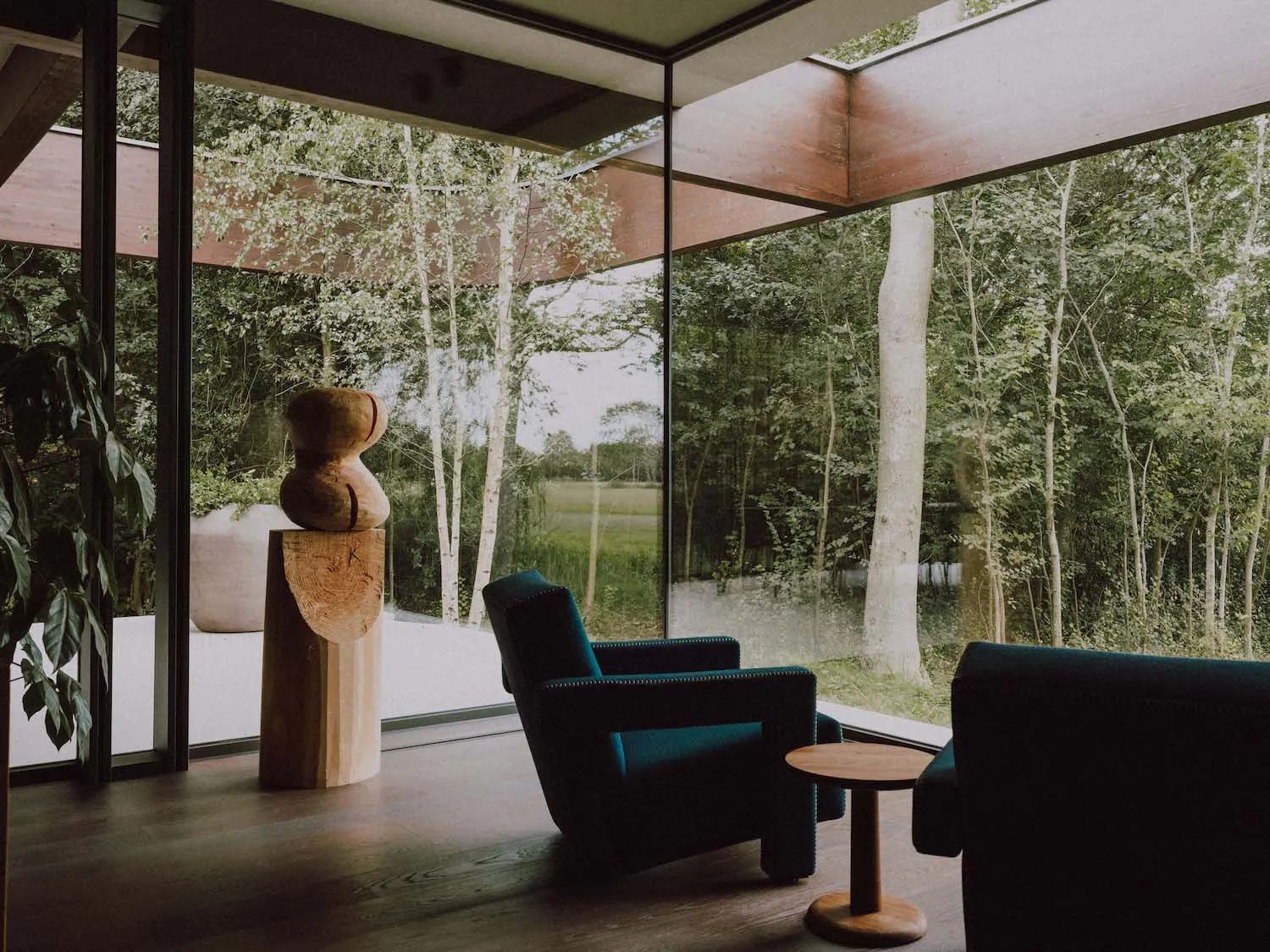 Modern living room interior featuring two teal upholstered armchairs, a small round wooden side table, a large abstract wooden sculpture on a pedestal, and expansive glass walls overlooking a lush green forest with birch trees.