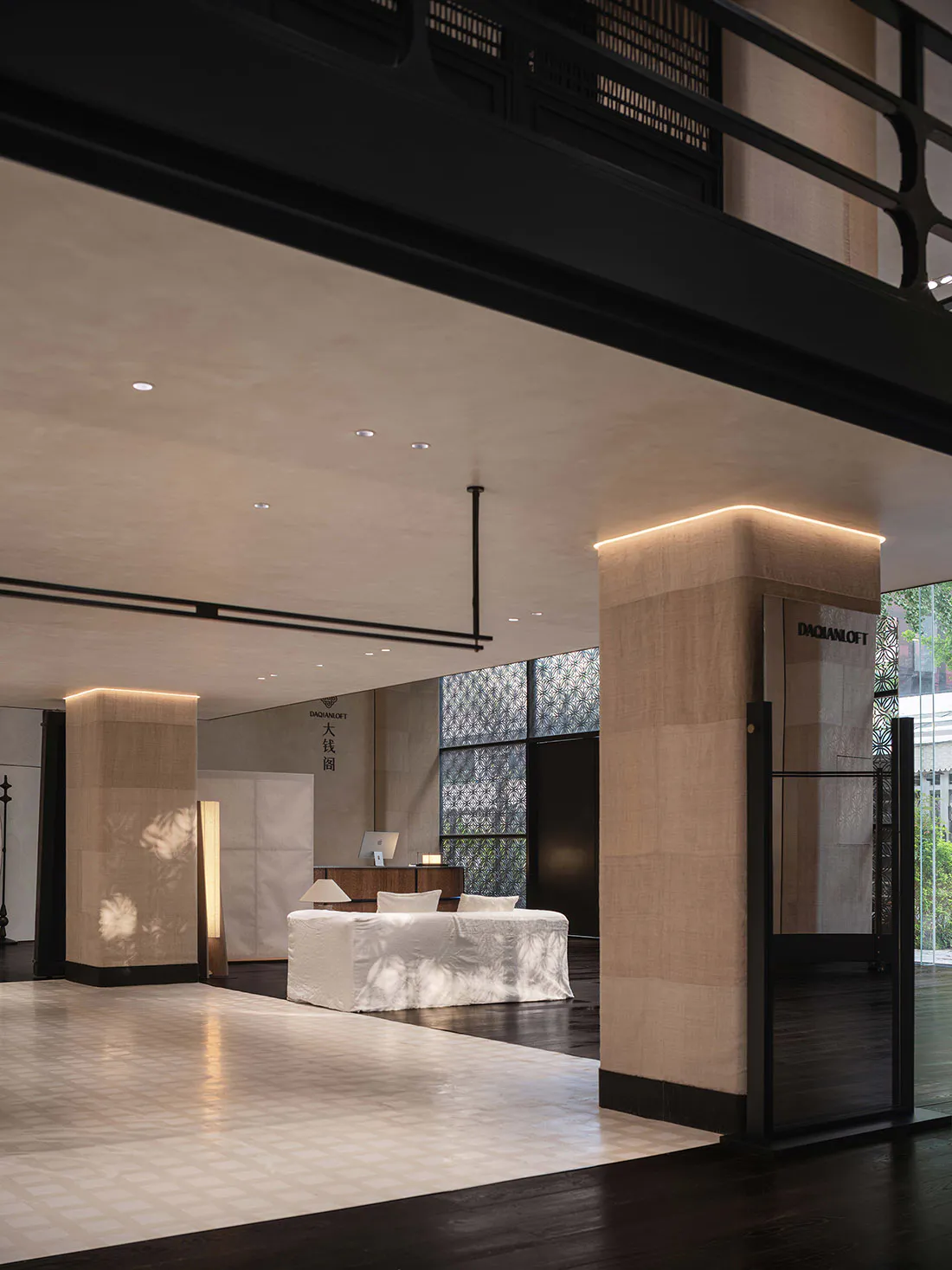 Modern minimalist reception area with tall beige linen-textured columns, white daybed, dark wood floors, and large patterned windows allowing dappled sunlight.