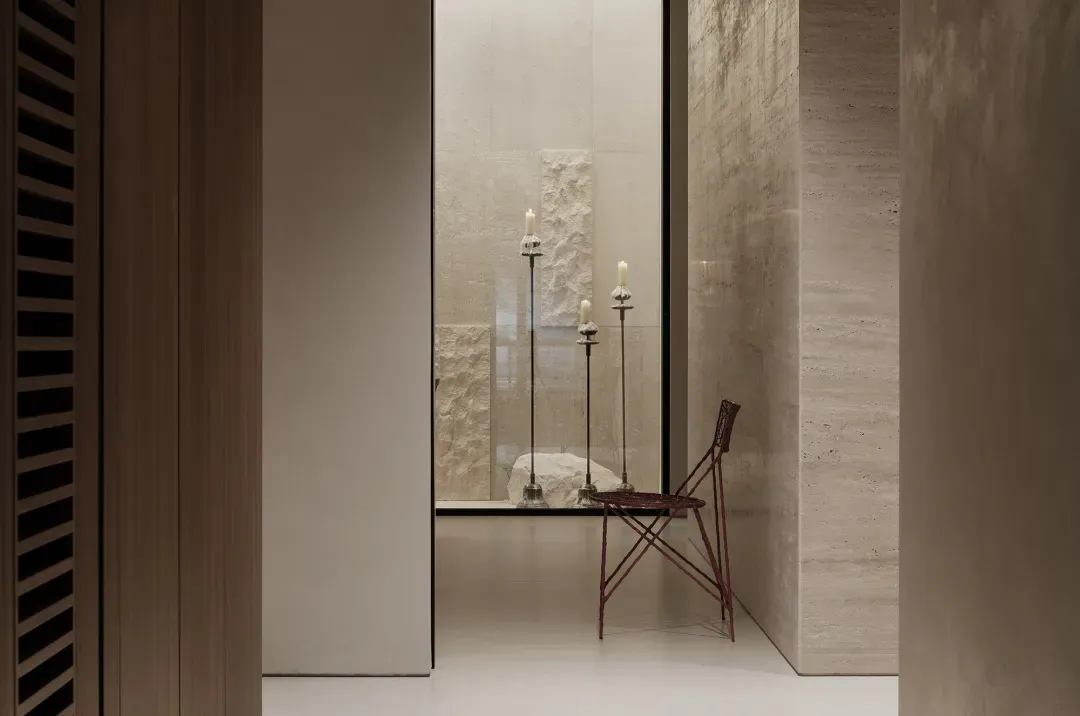 Minimalist interior view of a travertine-clad alcove featuring three tall candlesticks with lit candles, a large rough stone sculpture, and a distinctive red wireframe folding chair positioned against textured stone walls.