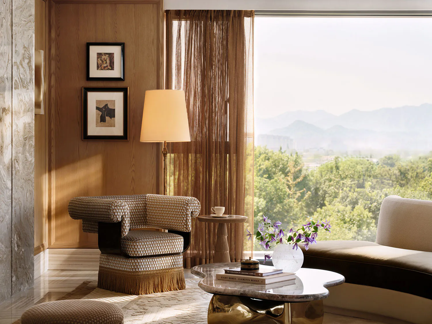 Elegant living room corner with walnut wood paneling, a patterned fringed lounge chair, marble coffee table, and large window overlooking green mountains.
