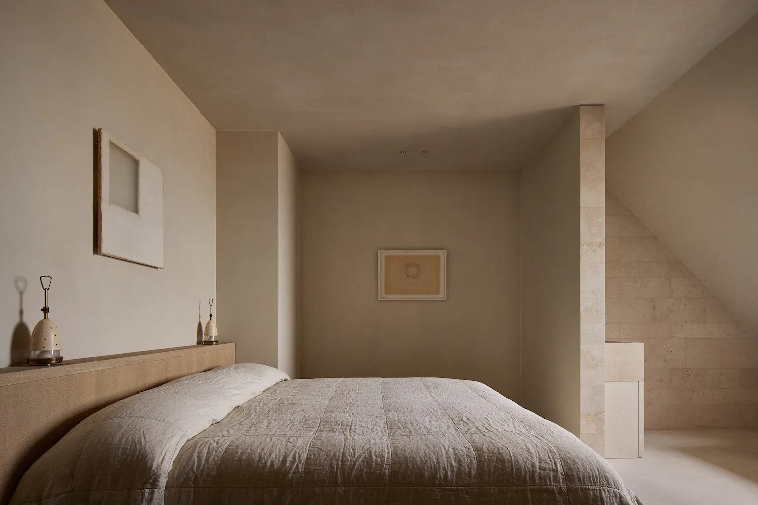 Minimalist bedroom interior with a large bed dressed in textured off-white linen, a wooden headboard shelf holding two ceramic lamps, travertine stone walls, a sloped ceiling, and framed abstract art on the walls.