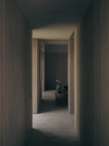Long narrow hallway with vertical wood paneling on walls and ceiling, concrete floor, and three modern leather chairs on a dark rug at the end.