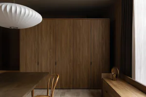 Modern minimalist dining area featuring a large walnut wood wardrobe, wooden dining table and chair, sculptural white pendant lamp, and a window seat with brass decor.