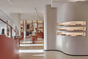 Modern minimalist retail boutique interior with curved white walls, terrazzo flooring, wooden display fixtures, and a prominent wall of illuminated shelves displaying sunglasses.