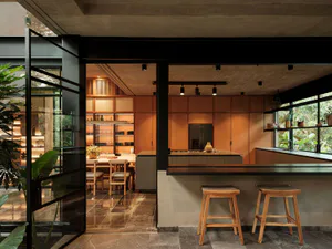 Modern open-plan kitchen featuring warm teak wood cabinetry, concrete island with wooden bar stools, dark tiled floor, and large openings revealing lush tropical greenery outside.